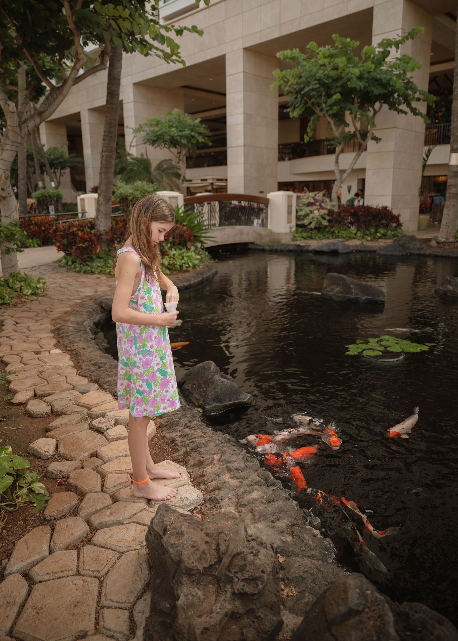 Young child in a floral dress stands by a koi pond with colorful fish at a tropical resort courtyard.