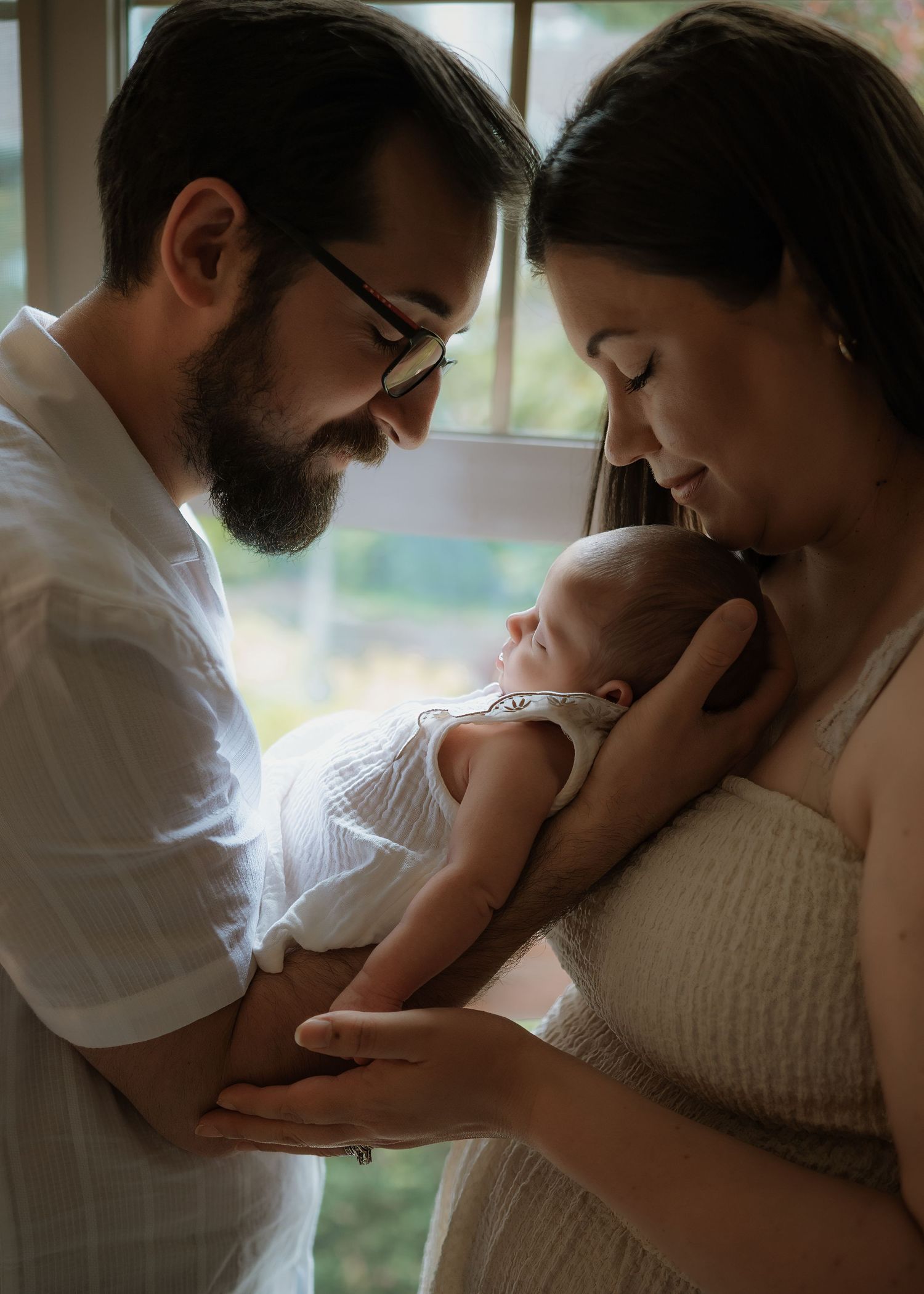 Bearded father with glasses gazes lovingly at a newborn baby held in the arms of the mother beside a sunlit window.