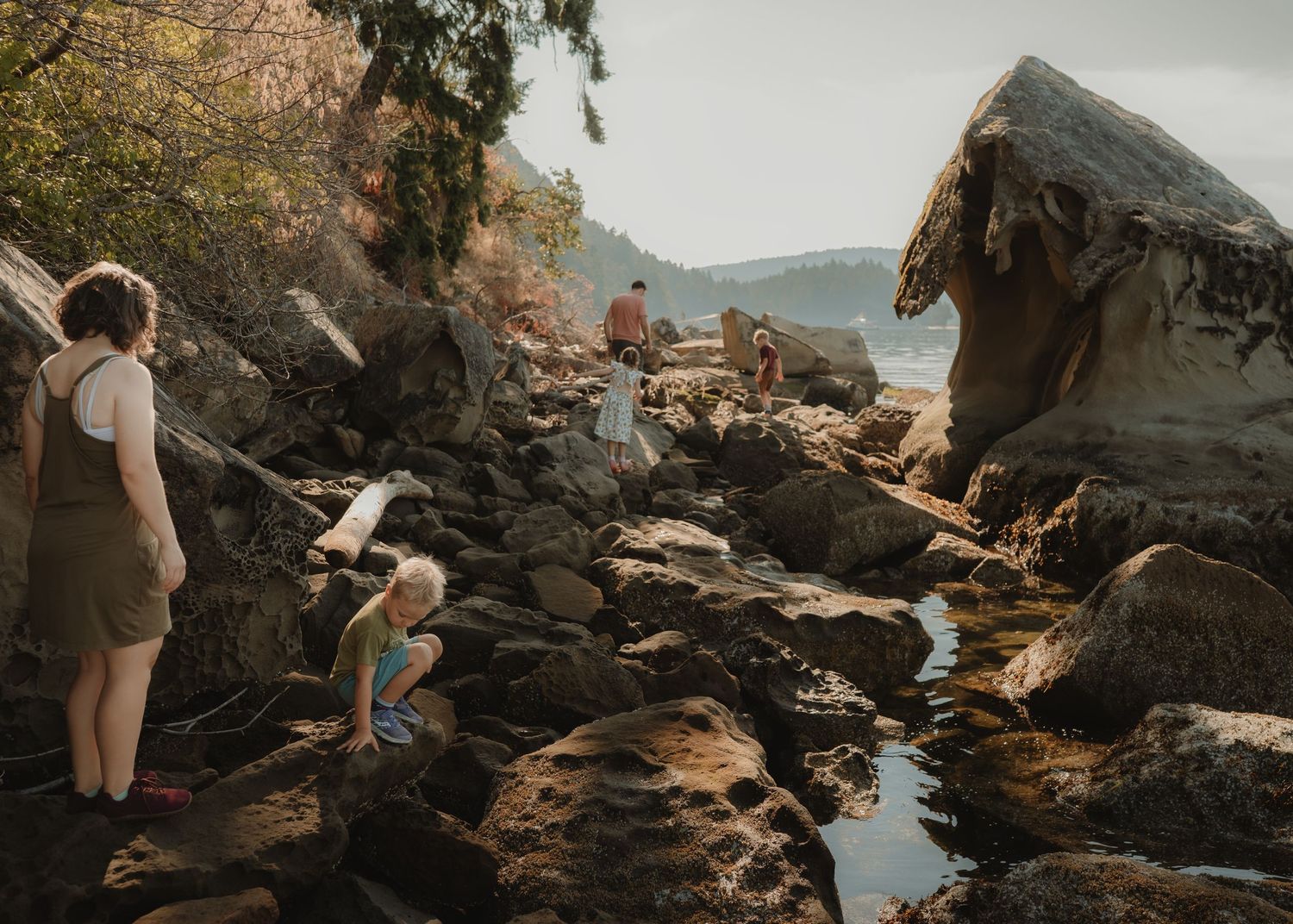 A woman stands on the rocky beach as a child crouches near a tide pool and a distant family explores sandstone formations.