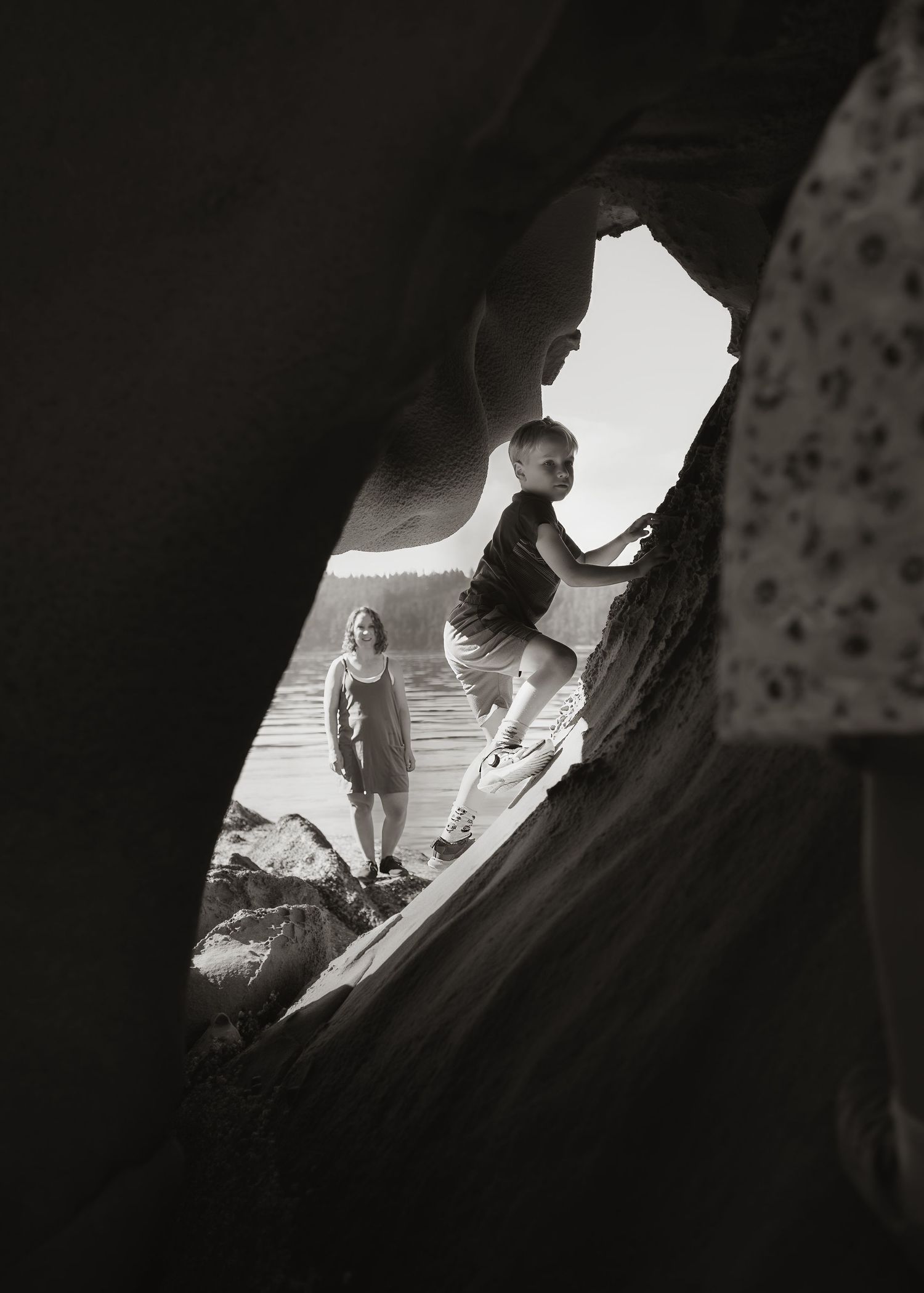 A black-and-white view through a rock opening shows a boy climbing sandstone while a woman watches from the shoreline.