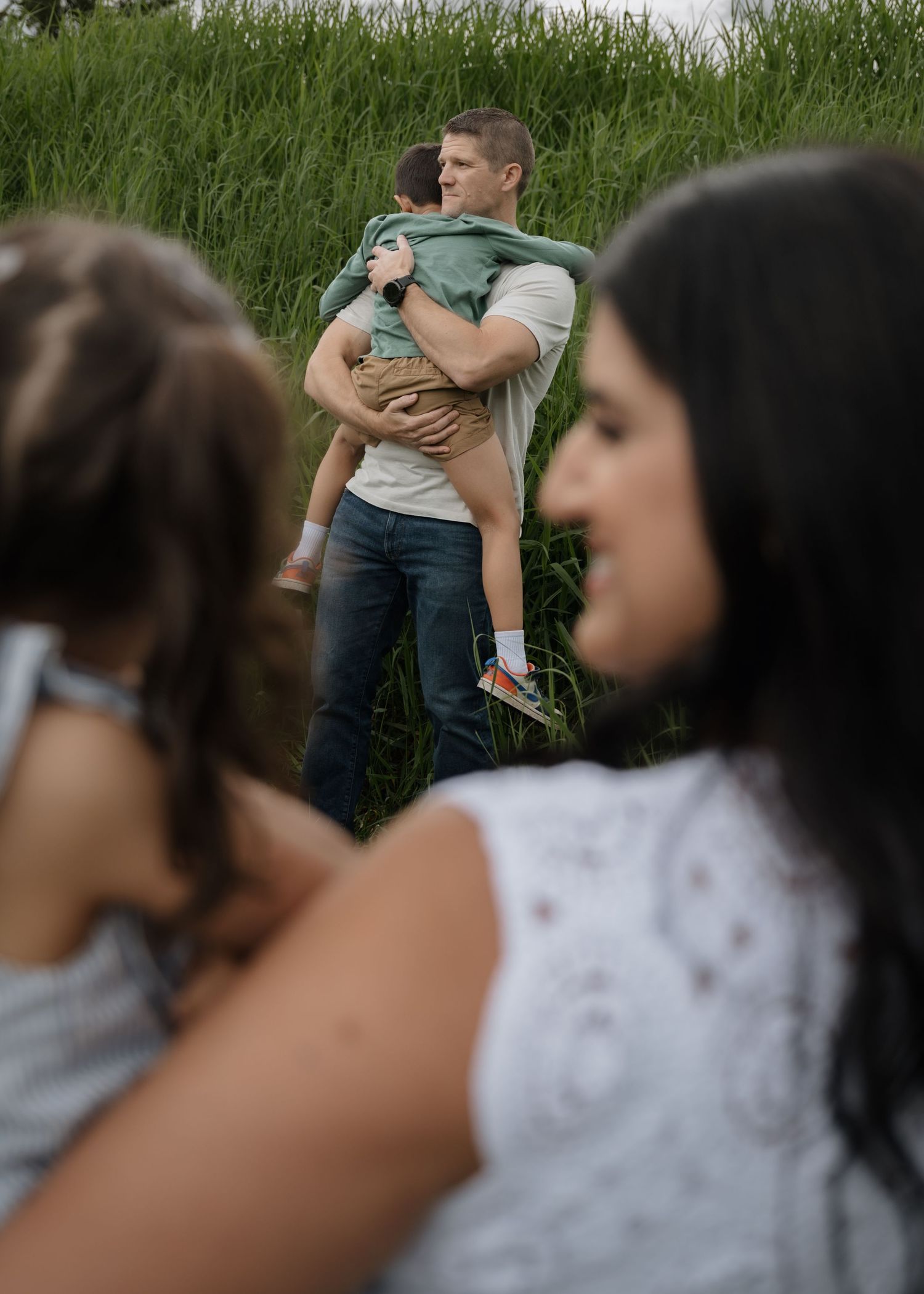 Man in a white shirt holds a child in a grassy field while two blurred family members stand in the foreground.