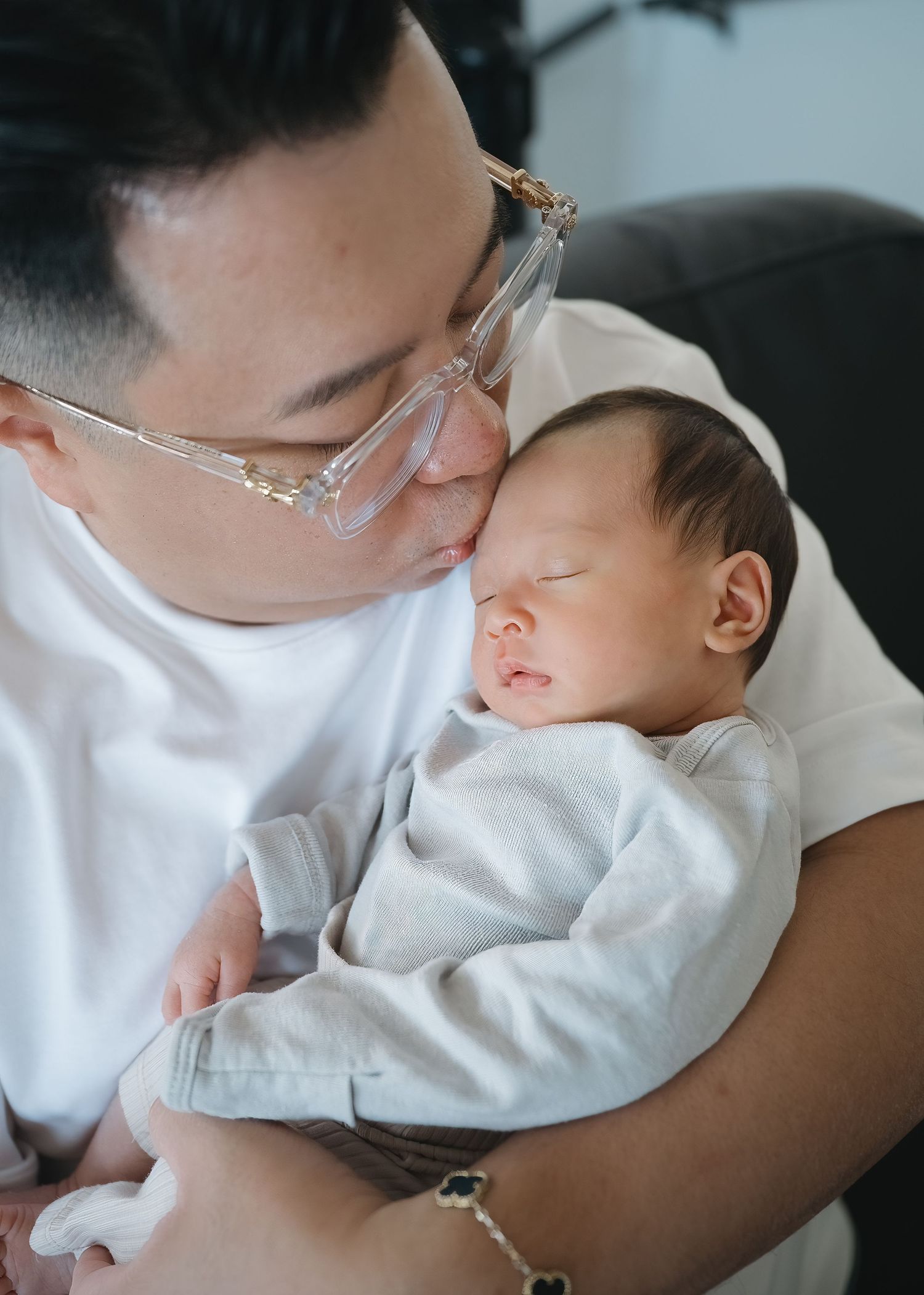 A father in glasses and a white t-shirt tenderly kisses the forehead of his sleeping newborn baby wrapped in a soft gray outfit.