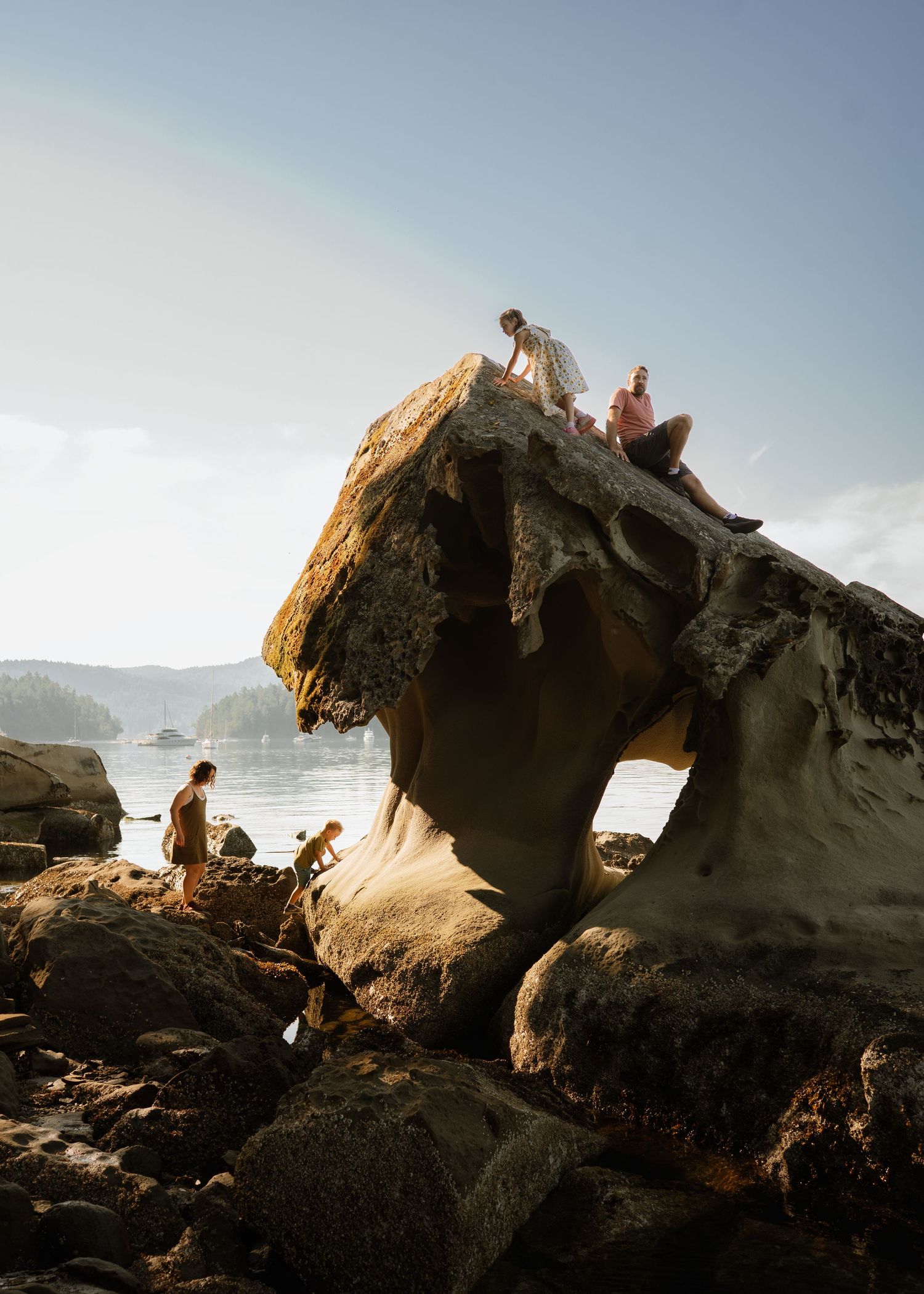 Two people sit atop a mushroom-shaped sandstone rock on a rocky shoreline while others explore below beside calm coastal waters.