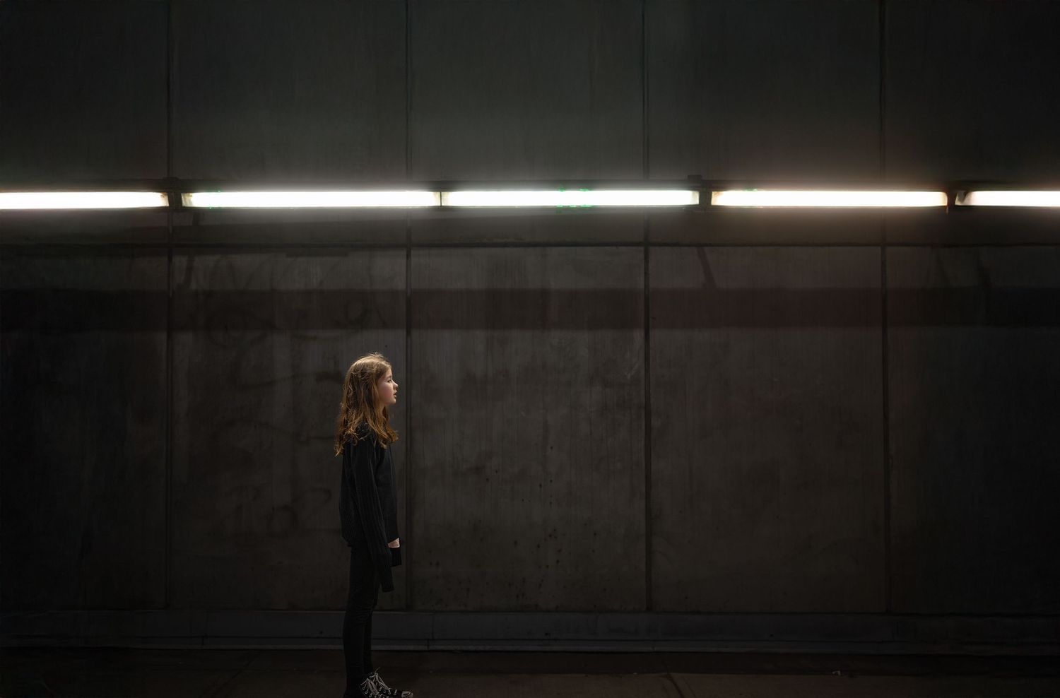 A silhouette standing alone in a dark concrete hallway illuminated by overhead fluorescent lights.