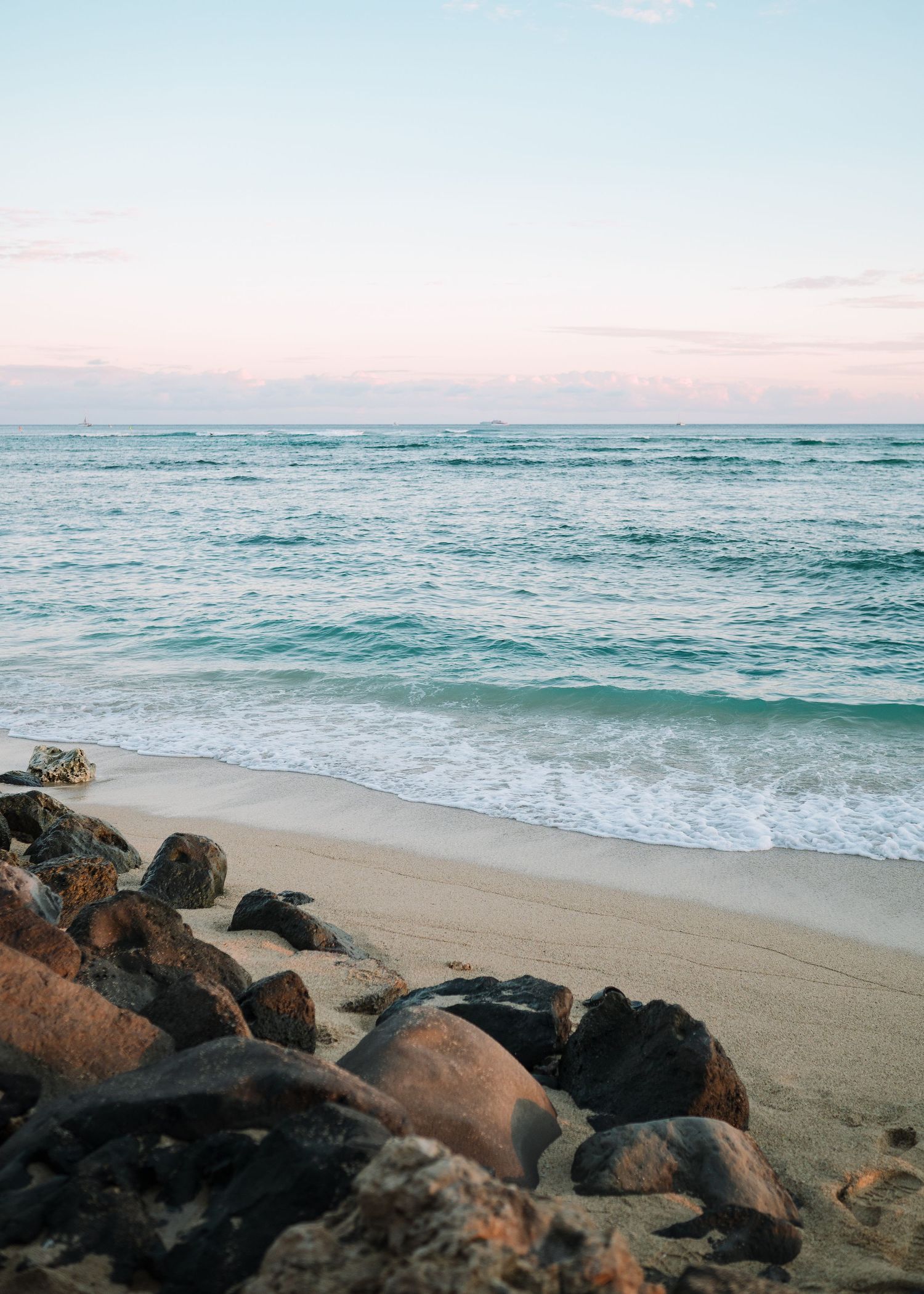 Turquoise waves roll onto a sandy beach lined with black volcanic rocks at sunset.