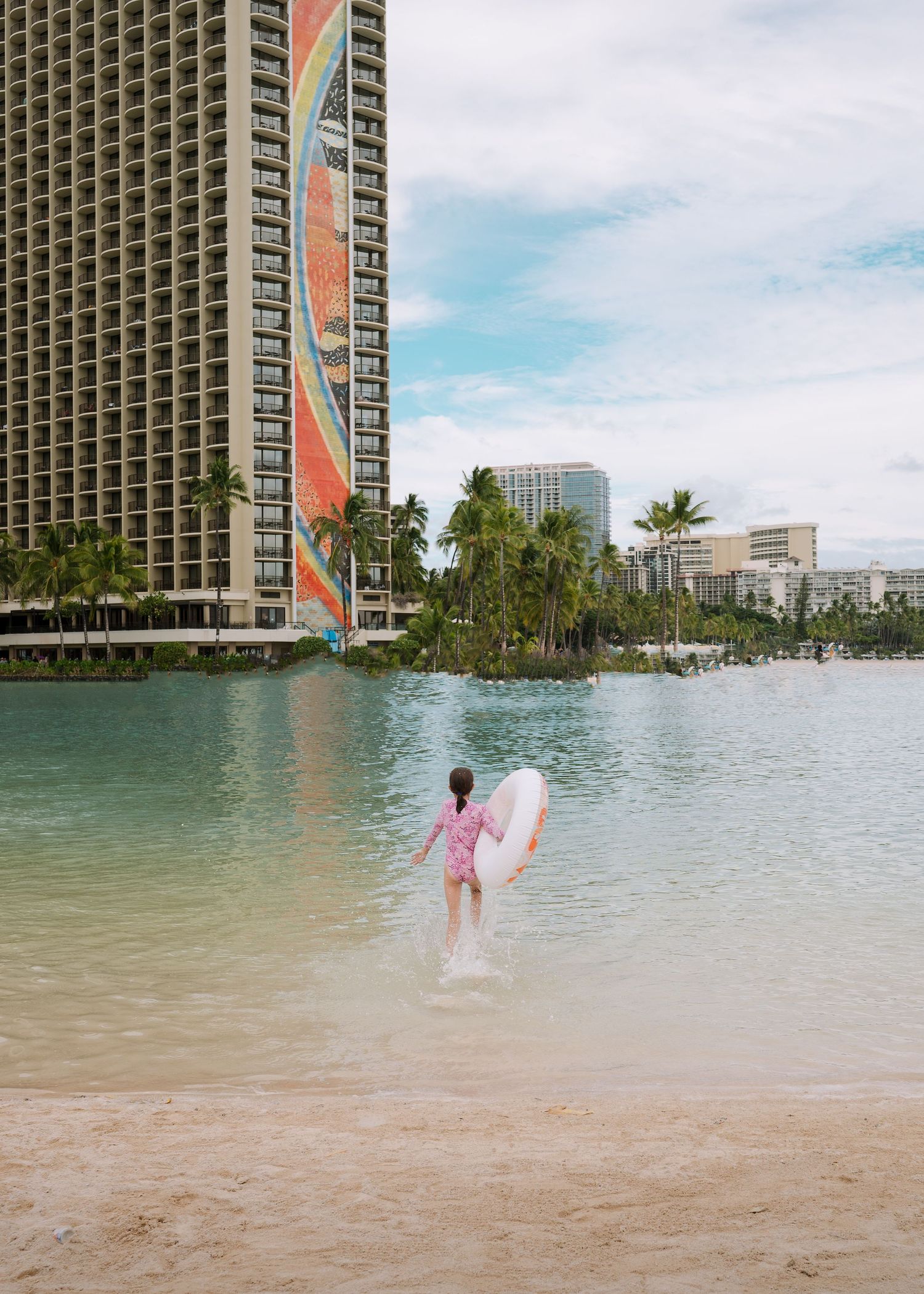 A surfer in pink walks into the lagoon waters near a high-rise hotel in Hawaii.