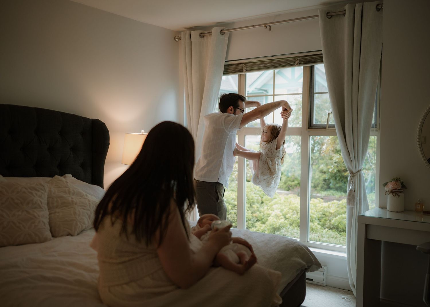 A father lifts a baby by the sunlit window while a mother sits on the bed holding another newborn in a cozy bedroom.