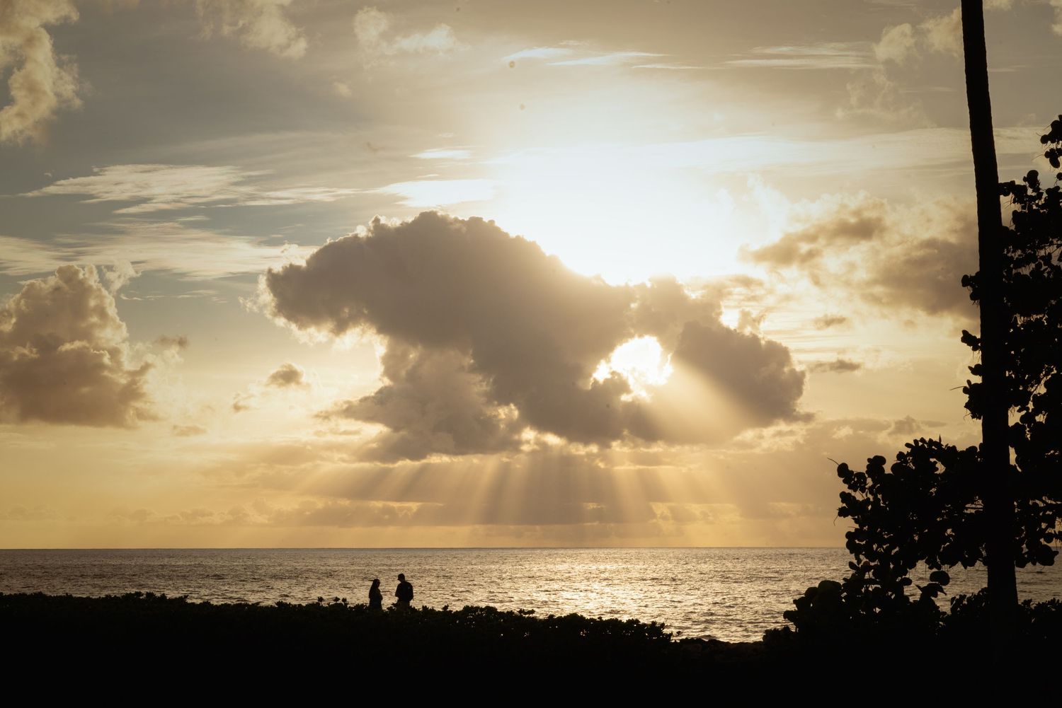 Silhouetted couple watches golden sunset rays burst through a large dark cloud over a calm ocean horizon.