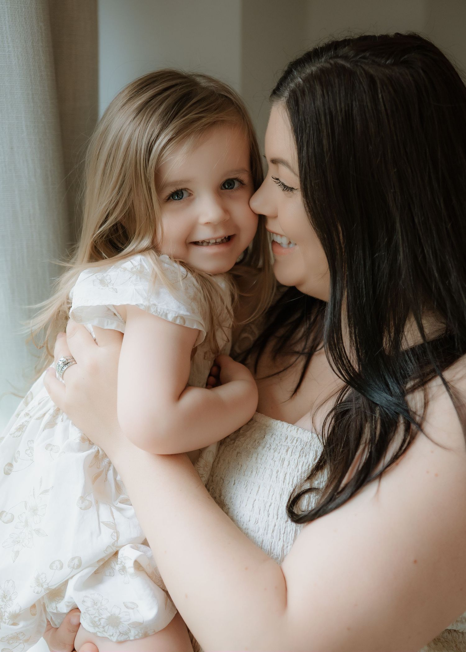 A smiling mother with long dark hair holds her toddler daughter in a floral dress, sharing a warm, affectionate moment together.