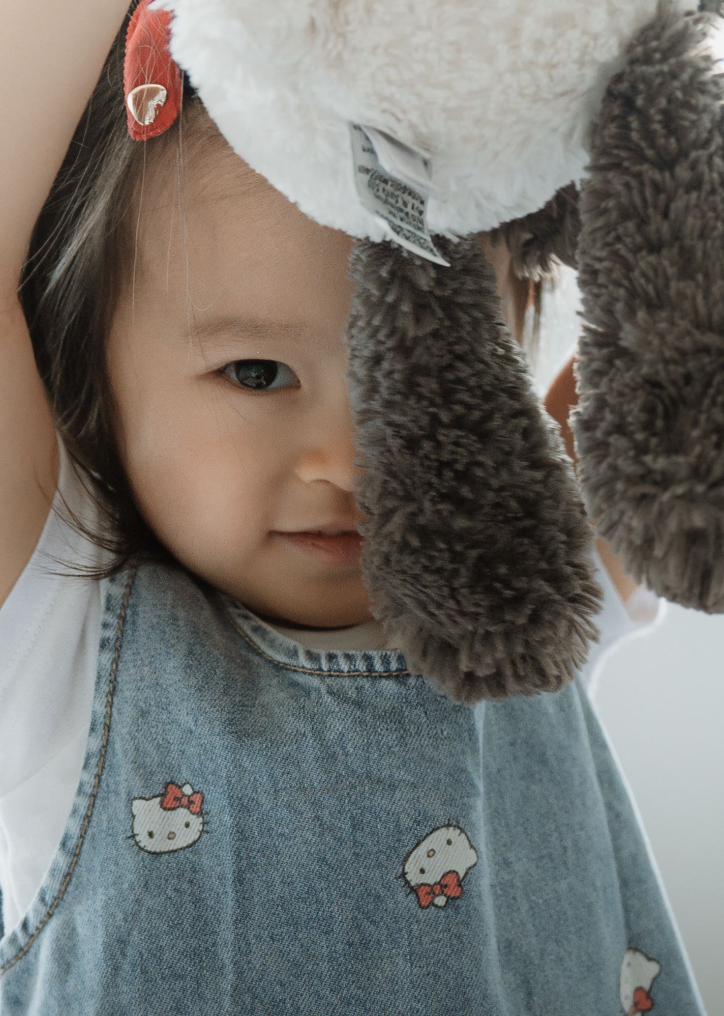 A smiling toddler in a Hello Kitty denim pinafore and red hair clip peeks out from behind a plush panda toy.