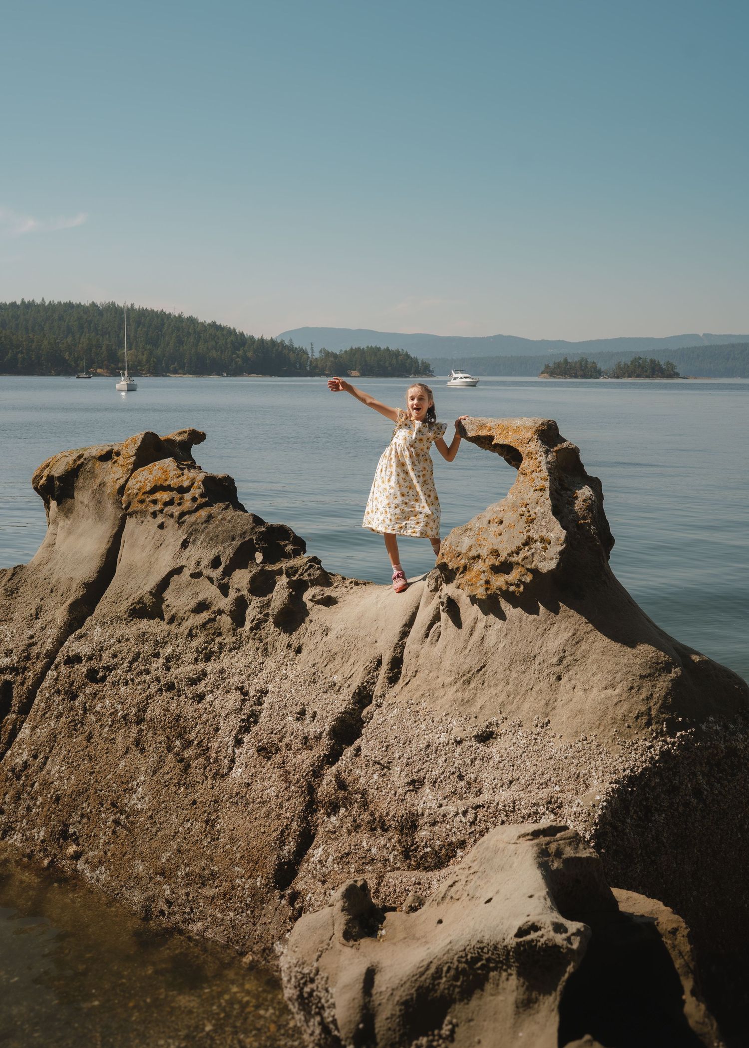 A girl in a white floral dress poses with outstretched arms on weathered sandstone rock above the sea and anchored sailboats.