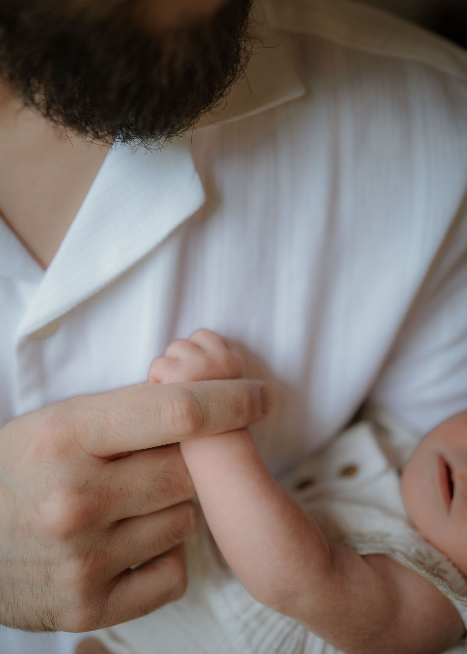 A newborn baby's tiny hand grasps a father's finger in a tender close-up of a bearded dad in a white linen shirt.