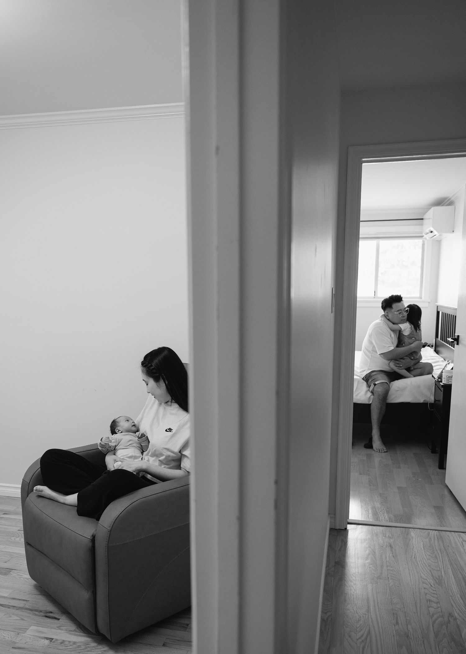Black and white doorway view of a mother holding her newborn in a chair while a father and child embrace on a bed in the next room.
