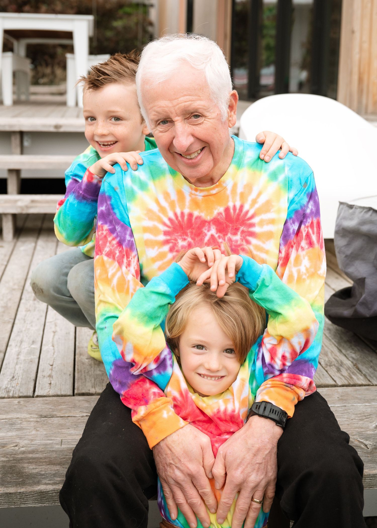 Smiling grandfather in a rainbow tie-dye sweatshirt sits on a wooden deck hugging two young grandsons in matching tie-dye shirts.