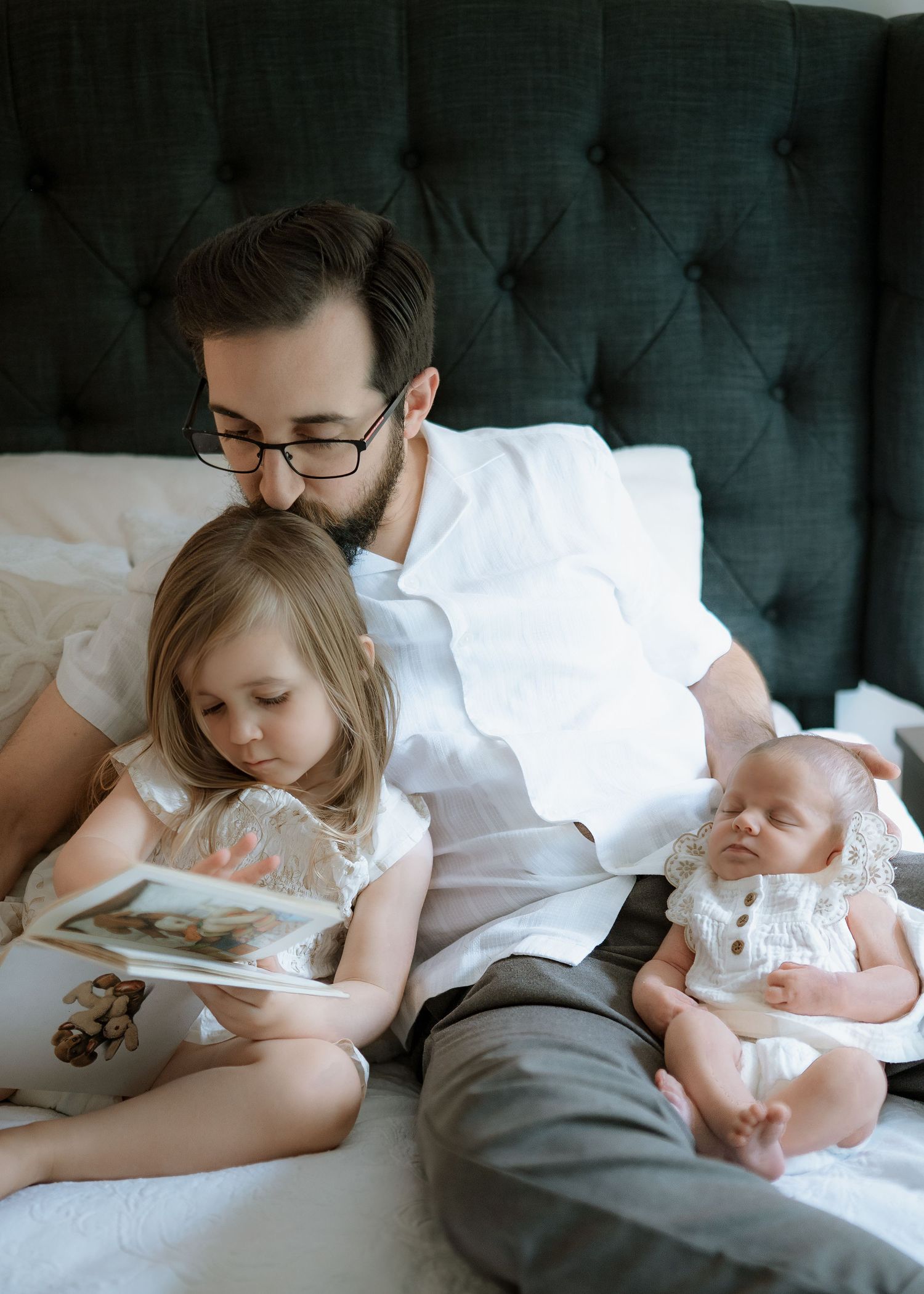 A bearded father in glasses sits on a bed reading a picture book with his toddler daughter while a newborn sleeps beside them.