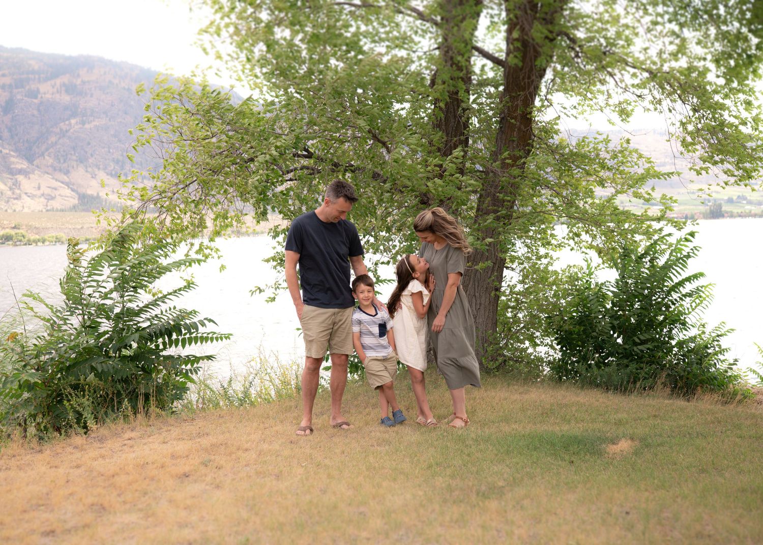 Family of four standing together on a grassy lakeside lawn under a large shady tree with mountains in the background.