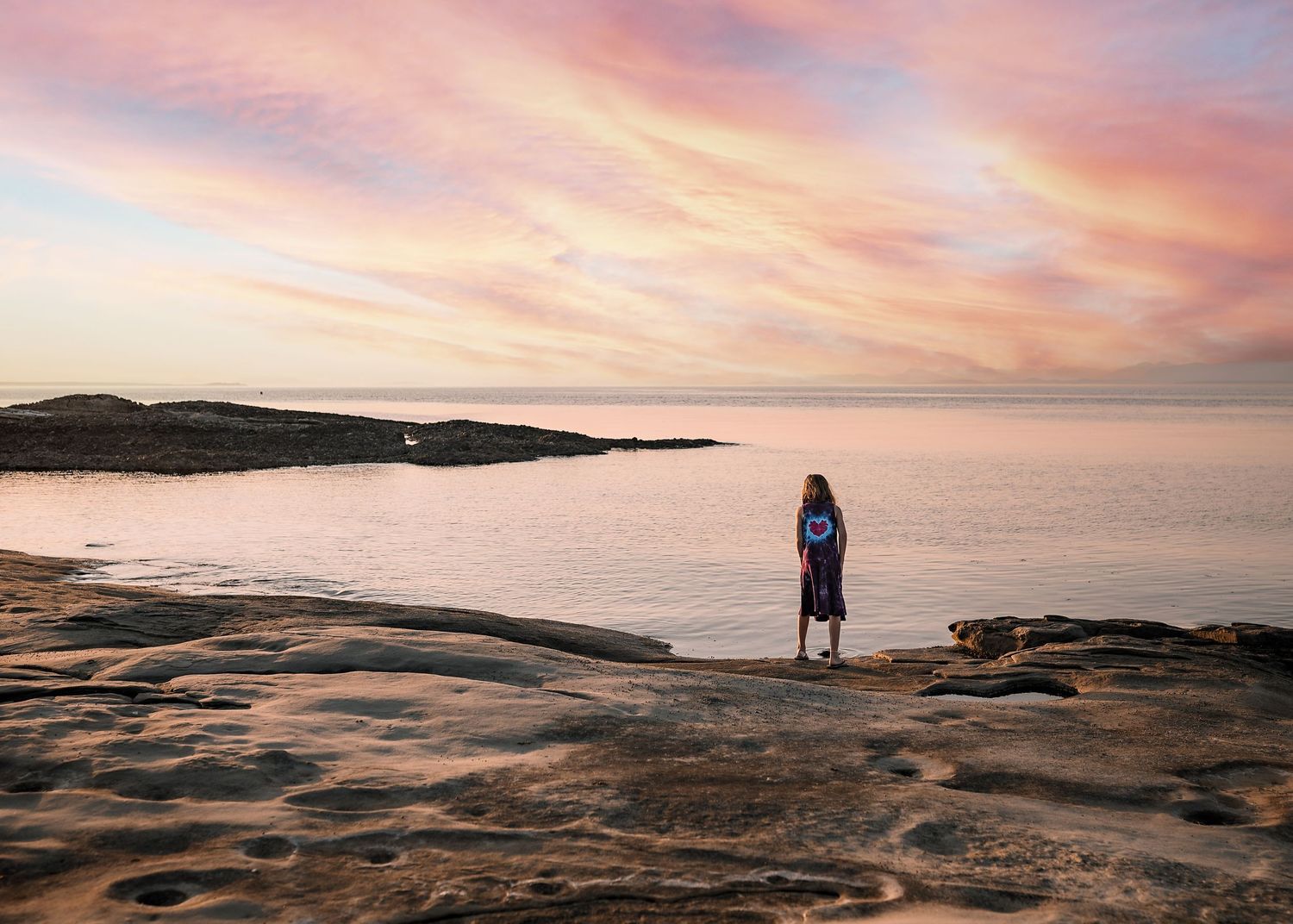 Person stands on smooth rocky shoreline gazing at a calm sea beneath a pink and orange sunset sky.