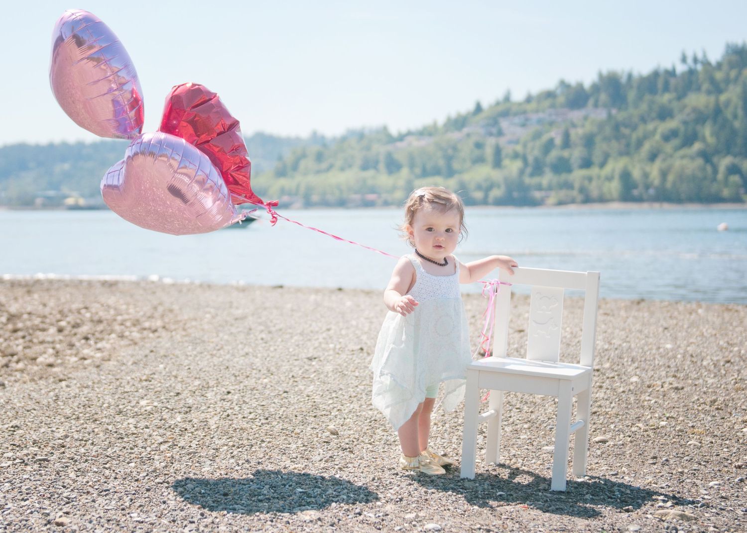 A toddler in a white dress stands on a beach holding pink balloons next to a white chair with water in the background.