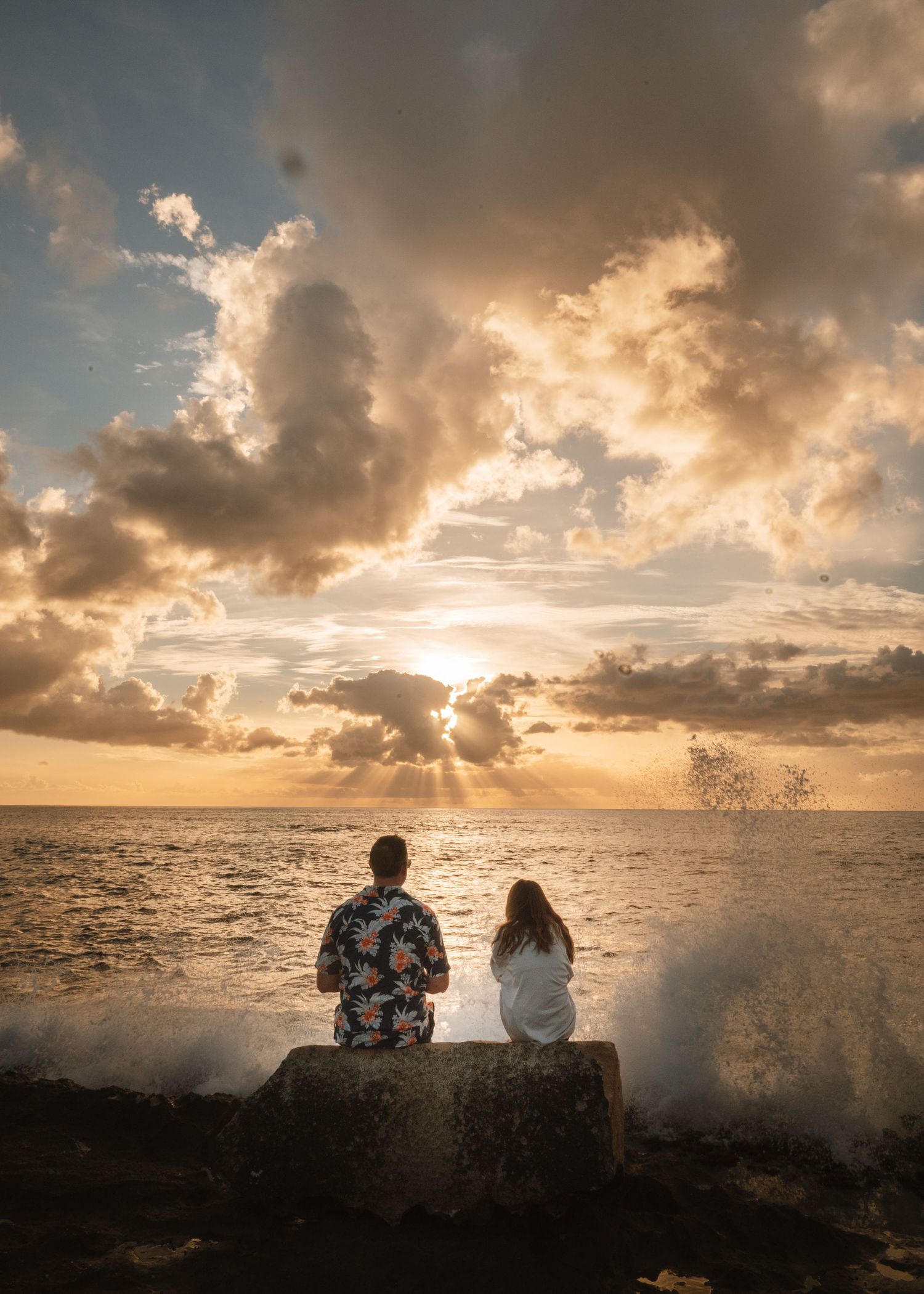 Silhouettes of people sitting on rocky seawall during dramatic golden sunset as waves crash against the shore.