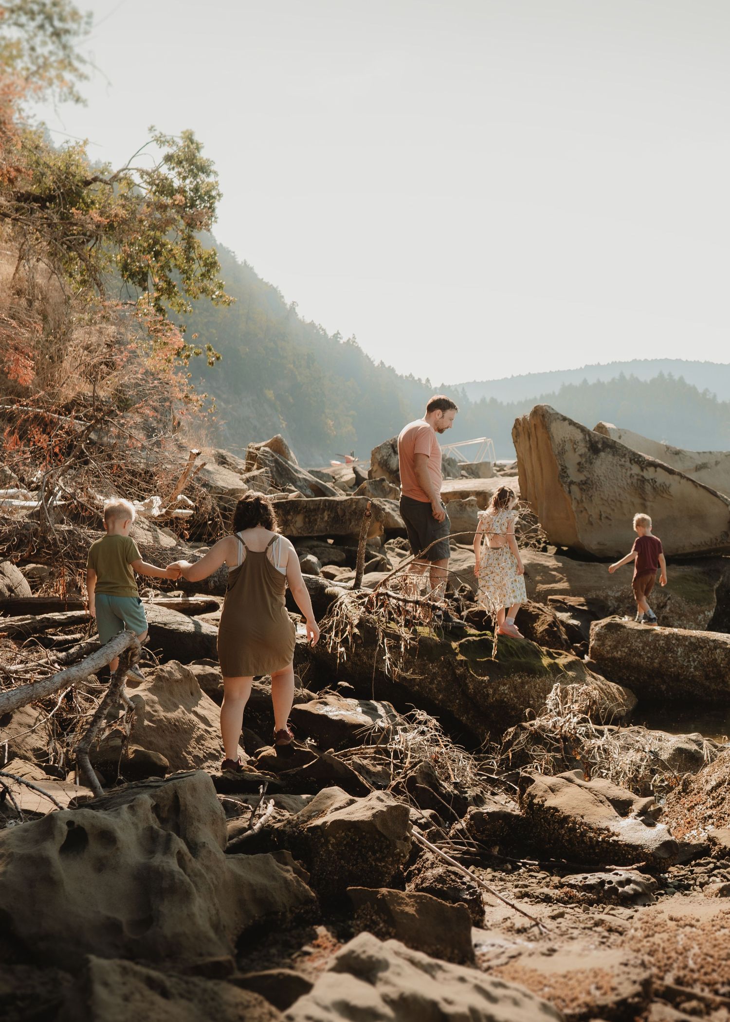 A family with children climbs and walks over rocks and driftwood on a rugged shoreline beneath forested hills.
