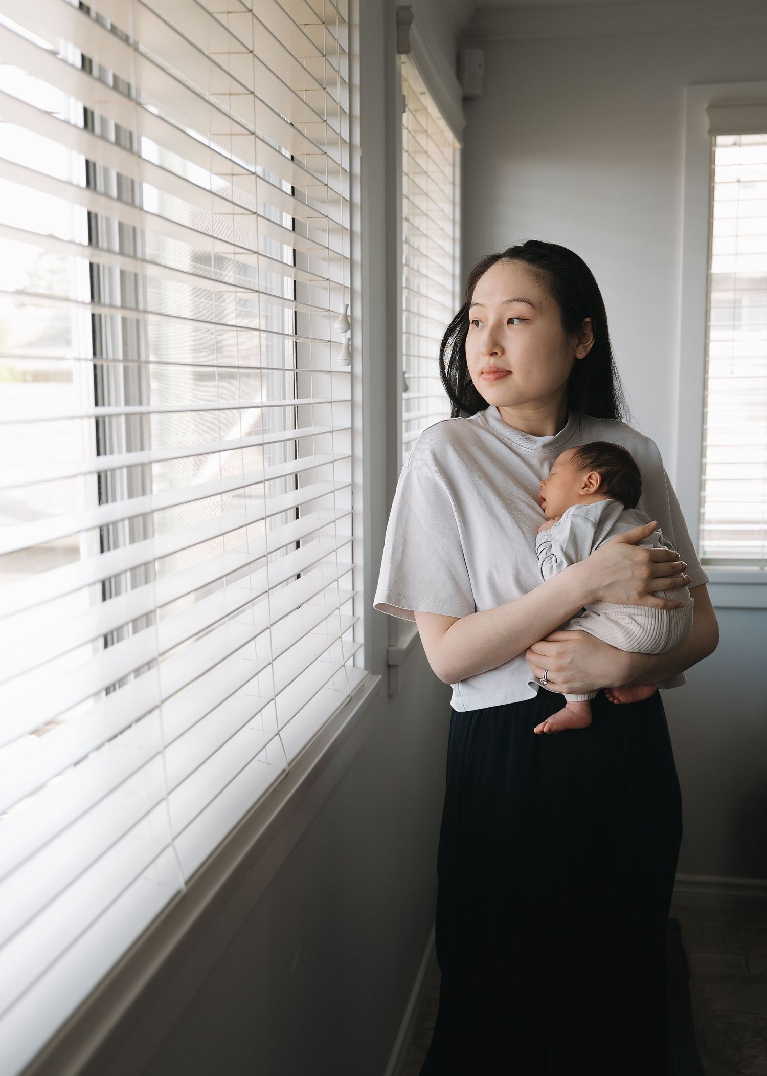 A mother in a gray top and black skirt holds her newborn while gazing out a window with white blinds in soft daylight.