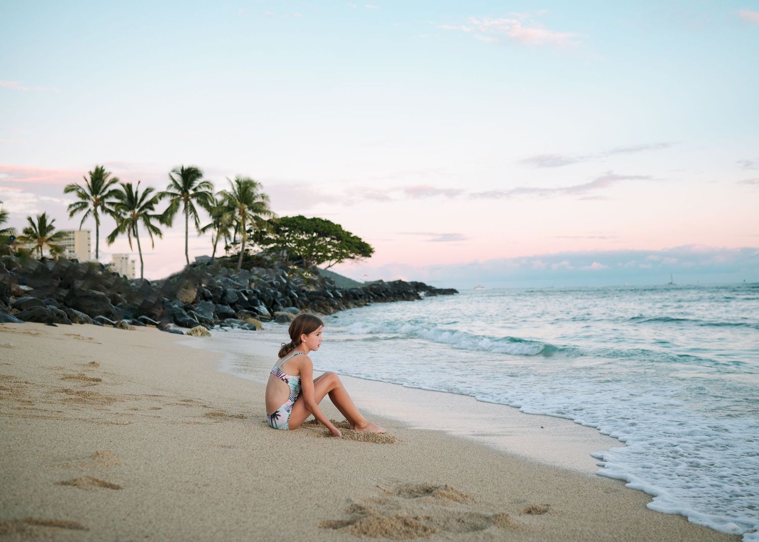 A sandy beach at sunset with palm trees and volcanic rocks along the shoreline.