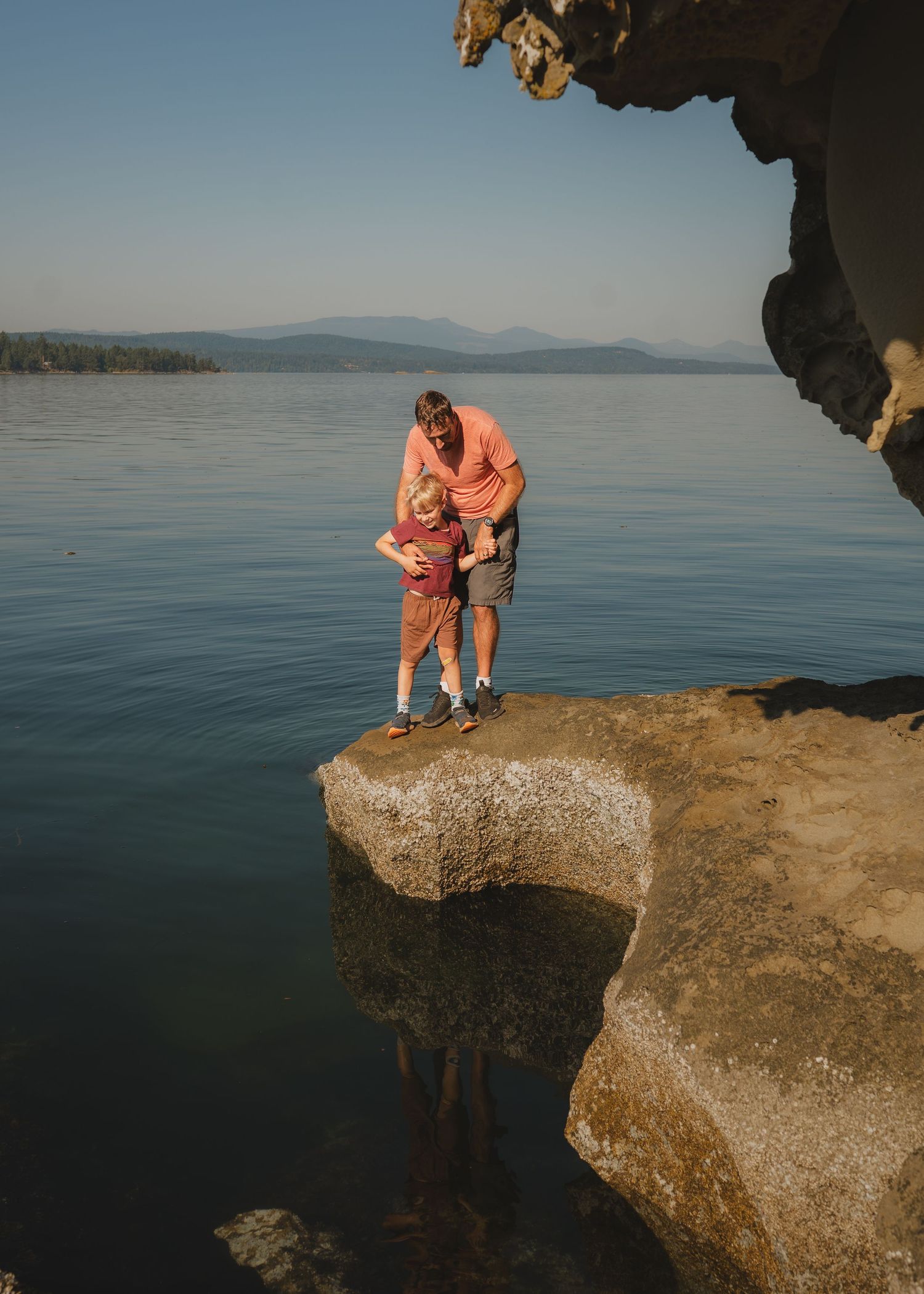 A father in an orange shirt holds a young child on a rocky ledge above calm blue water with distant hazy mountains.