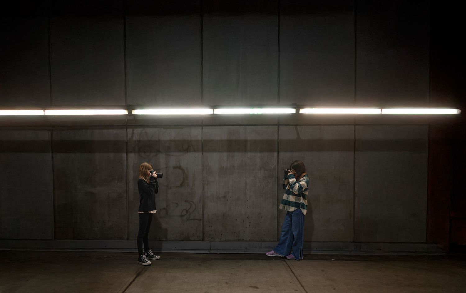 Two people stand apart under fluorescent lighting against a dark concrete wall in a minimalist urban setting.