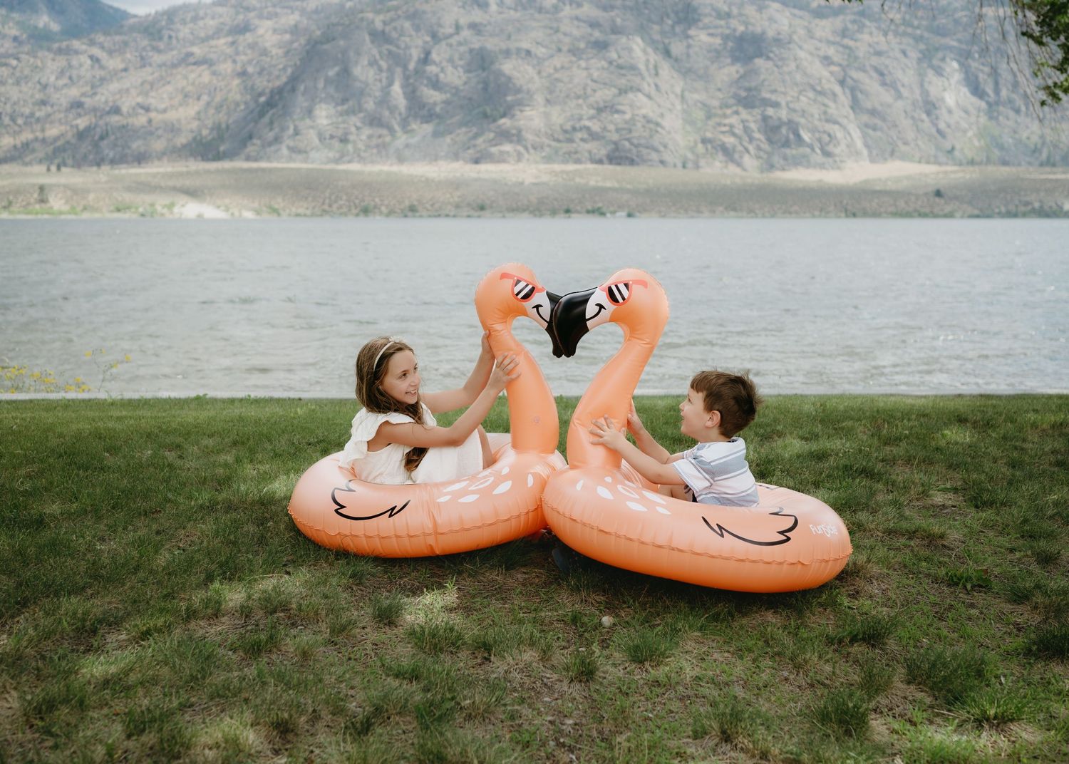 Two people lounge on peach-colored pool floats on grass by a serene lake with mountains in the background.
