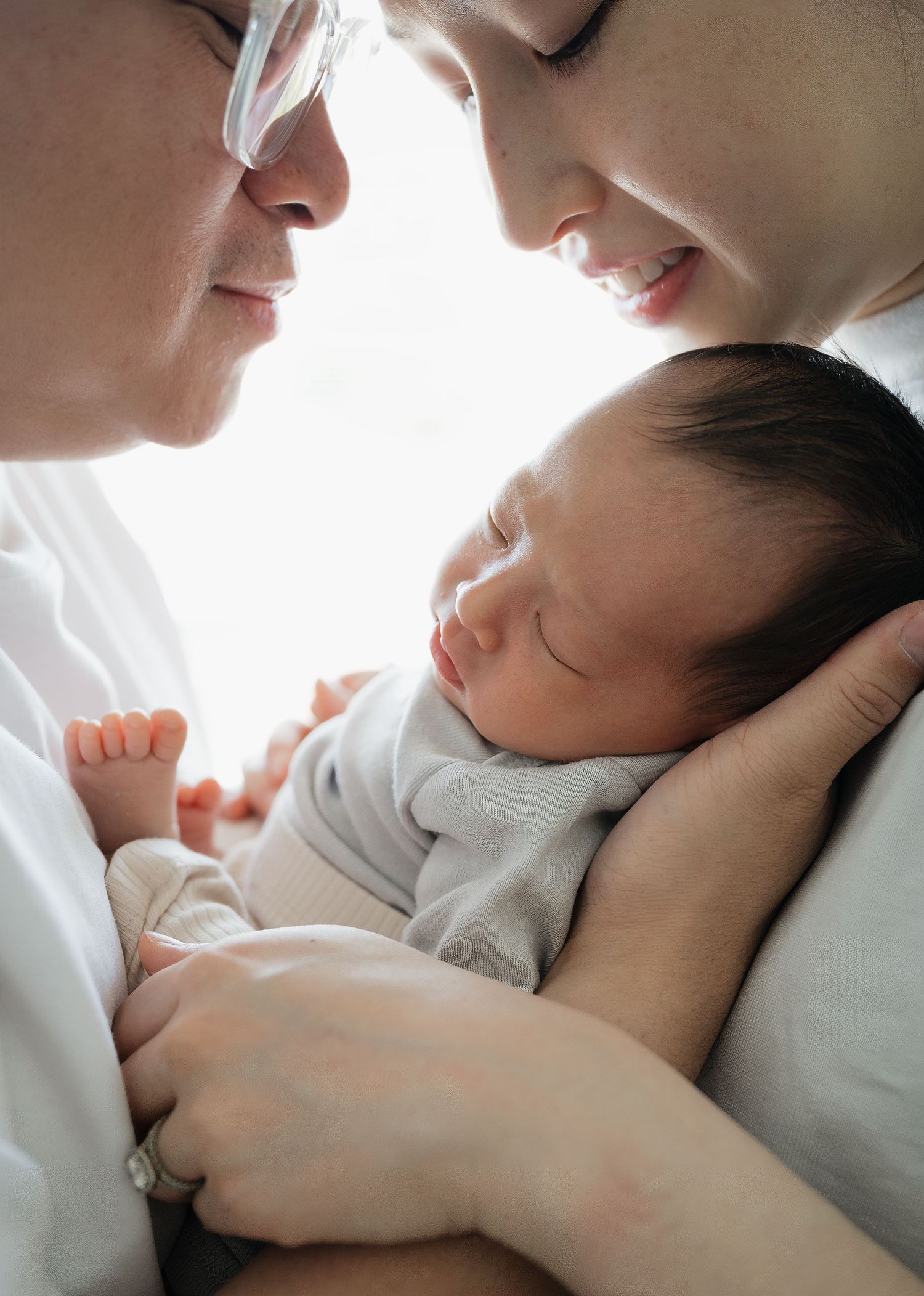 Two parents lean in close with their foreheads nearly touching as they cradle their sleeping newborn baby in bright natural light.