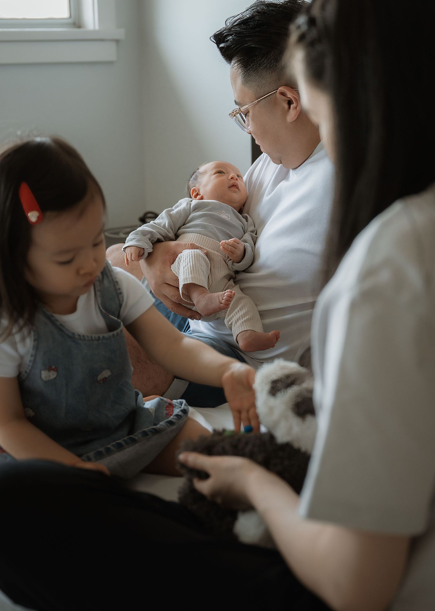 A family sits together on a bed as a father cradles his newborn, while a toddler girl and mother hold a plush toy nearby.
