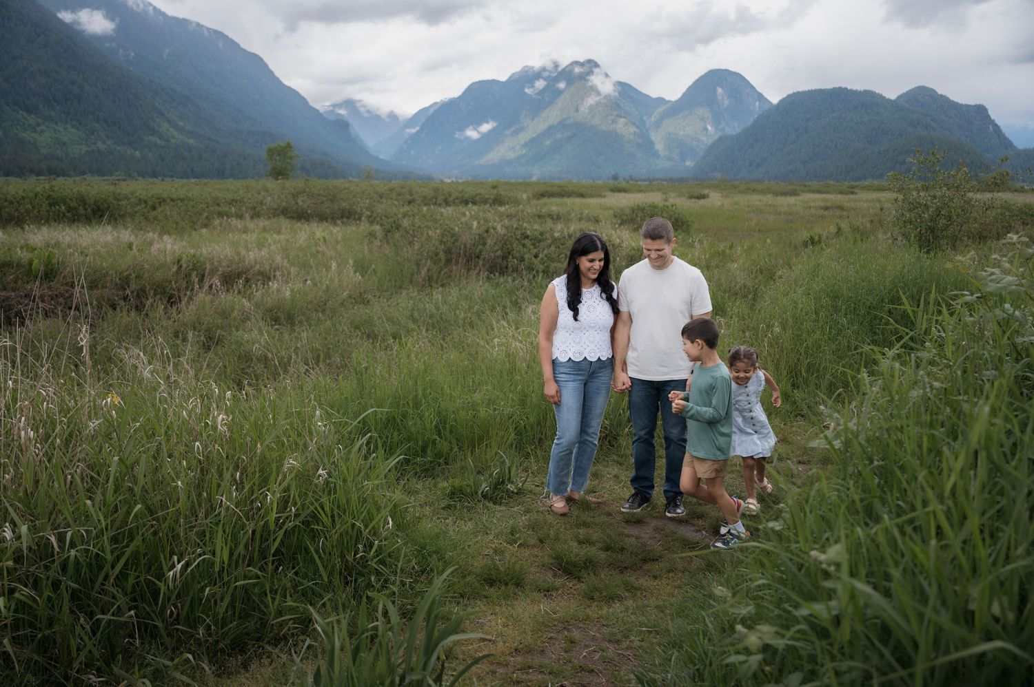 Family of four walking together through tall green grass in a wide valley with misty mountains in the background.