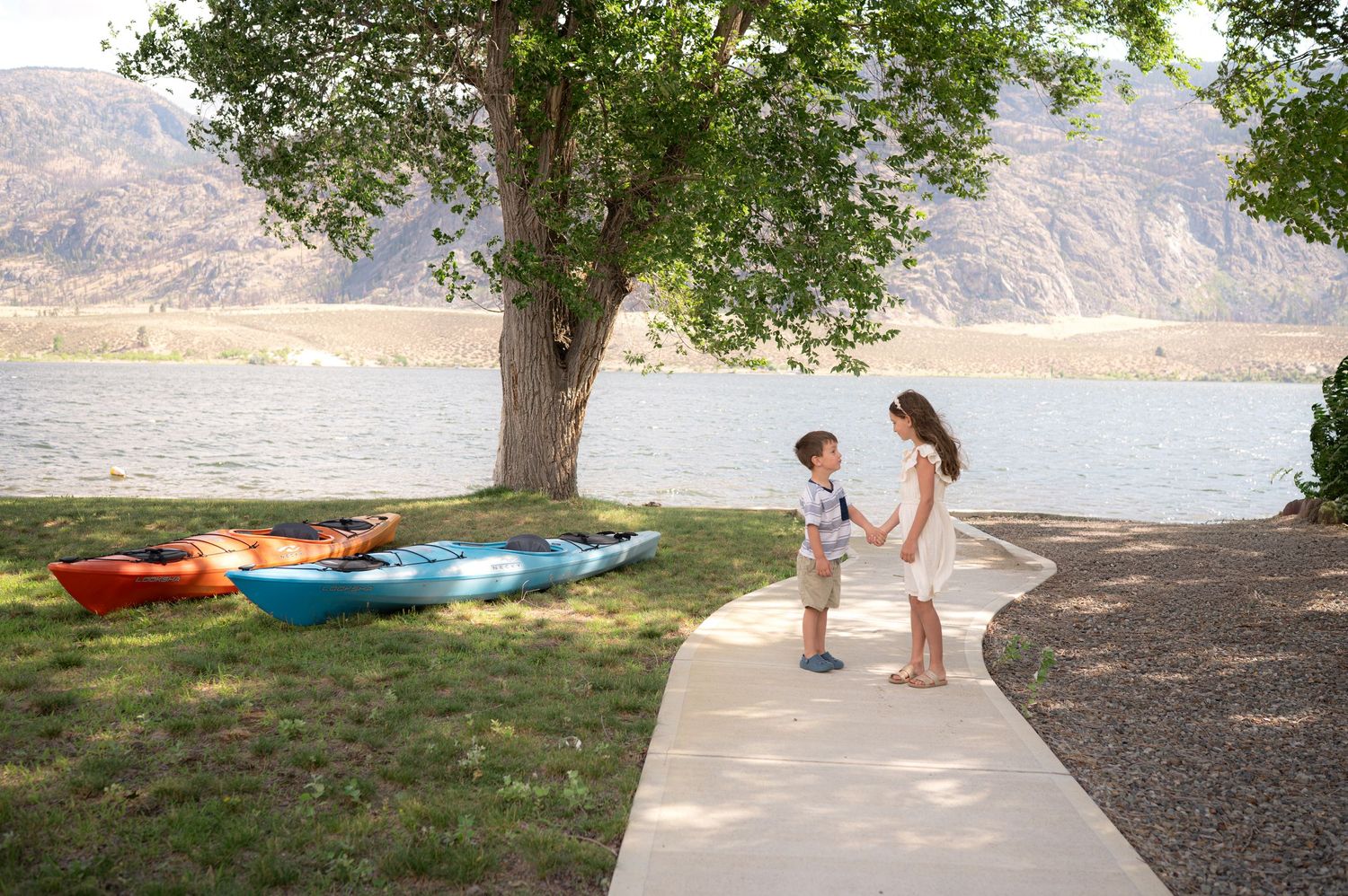 Two children hold hands on a sunny lakeside path near colorful kayaks resting on the grass beneath a large shade tree with mountains behind.