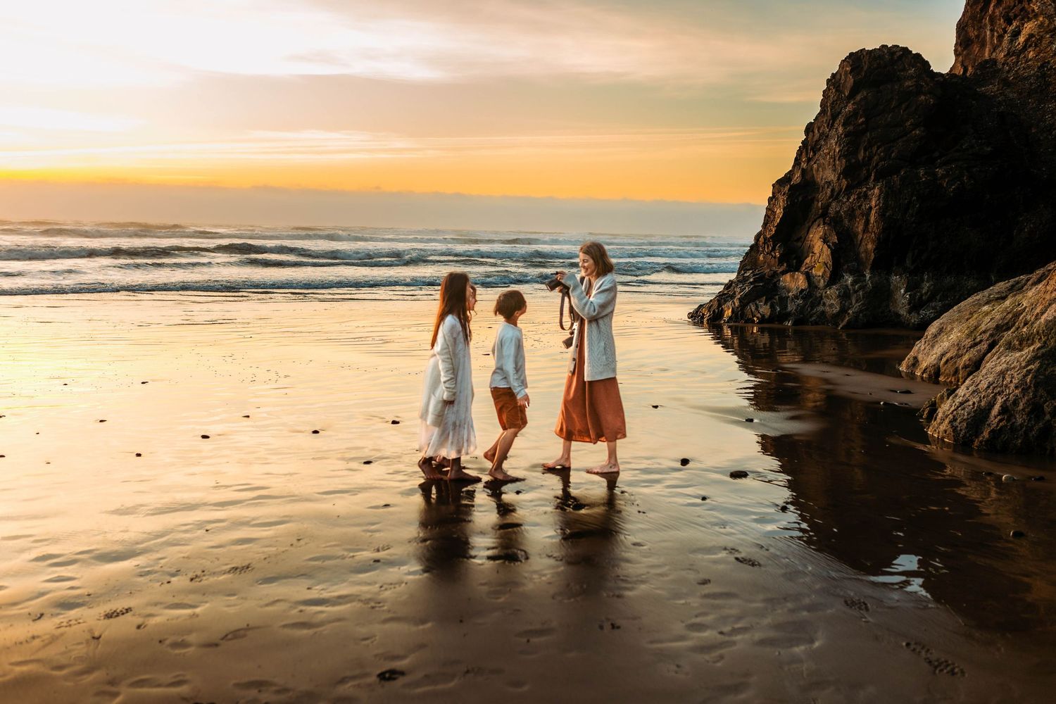 Mother and two children walk along a glossy beach at sunset with waves rolling in and dark rocks nearby.
