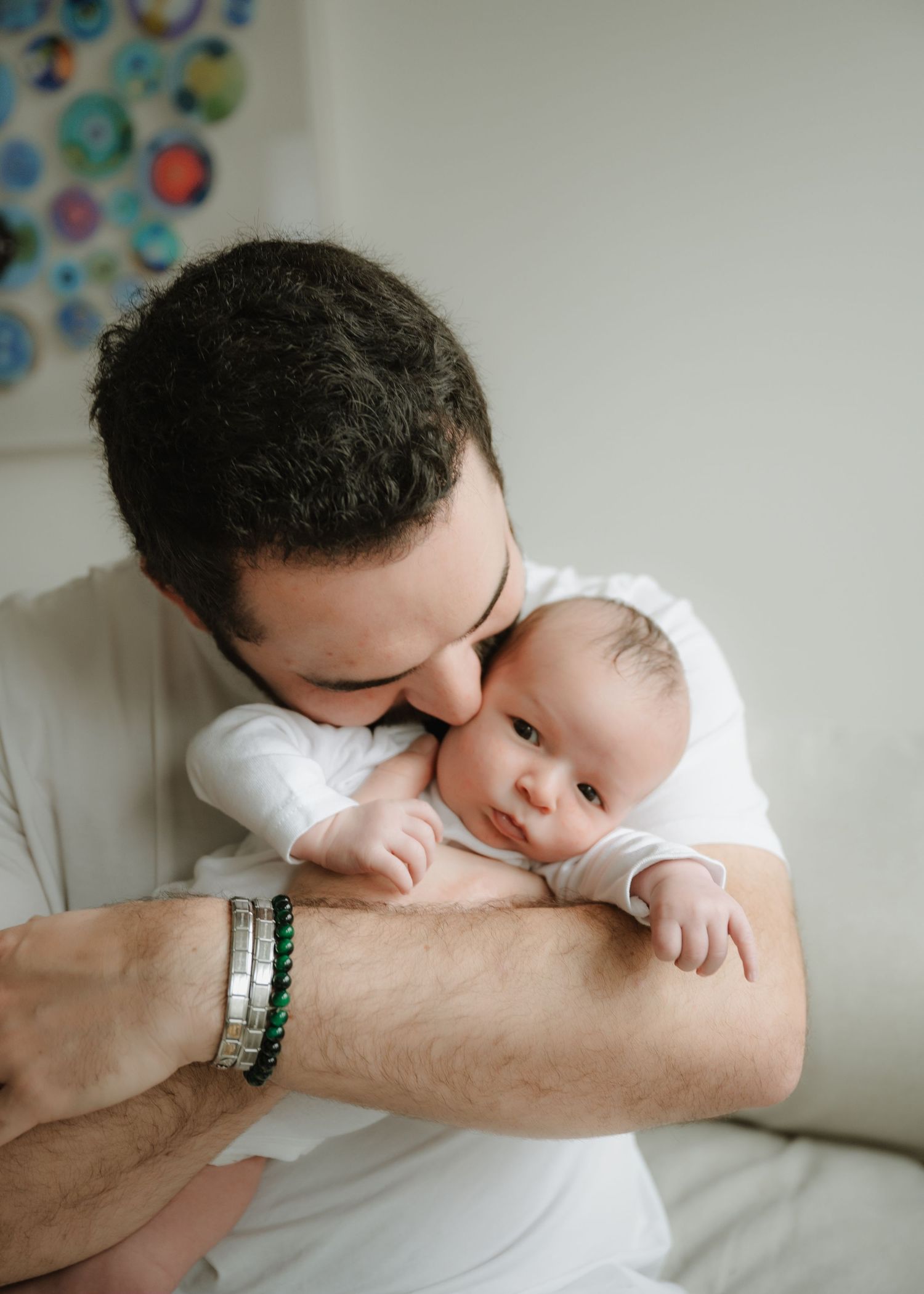 Father in a white shirt kisses his wide-eyed newborn baby's cheek while holding the infant close on a cozy couch.