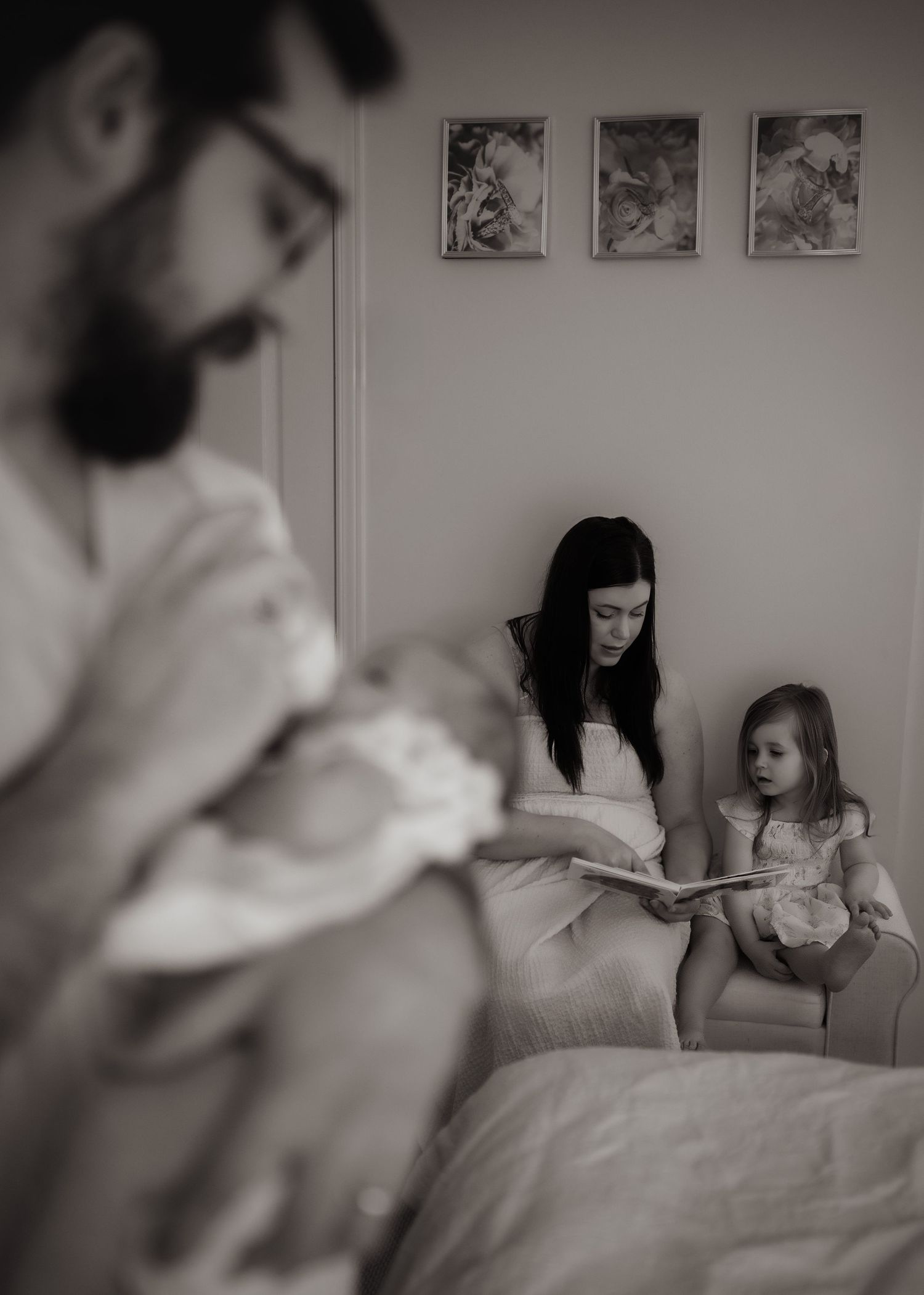 A mother reads a picture book to her young daughter while a bearded father in the foreground cradles a newborn baby.