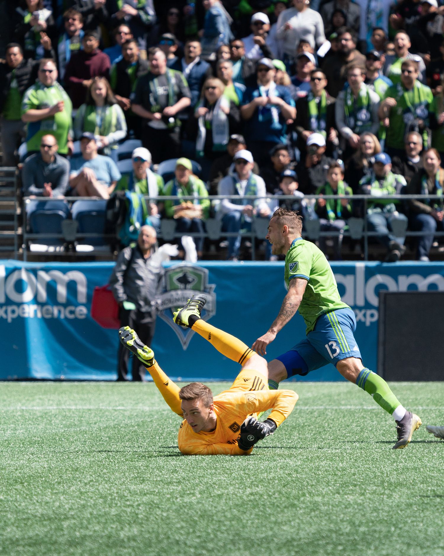 Soccer goalkeeper in orange falls to the grass as a green-jersey player runs past while fans in green fill the stadium stands behind them.
