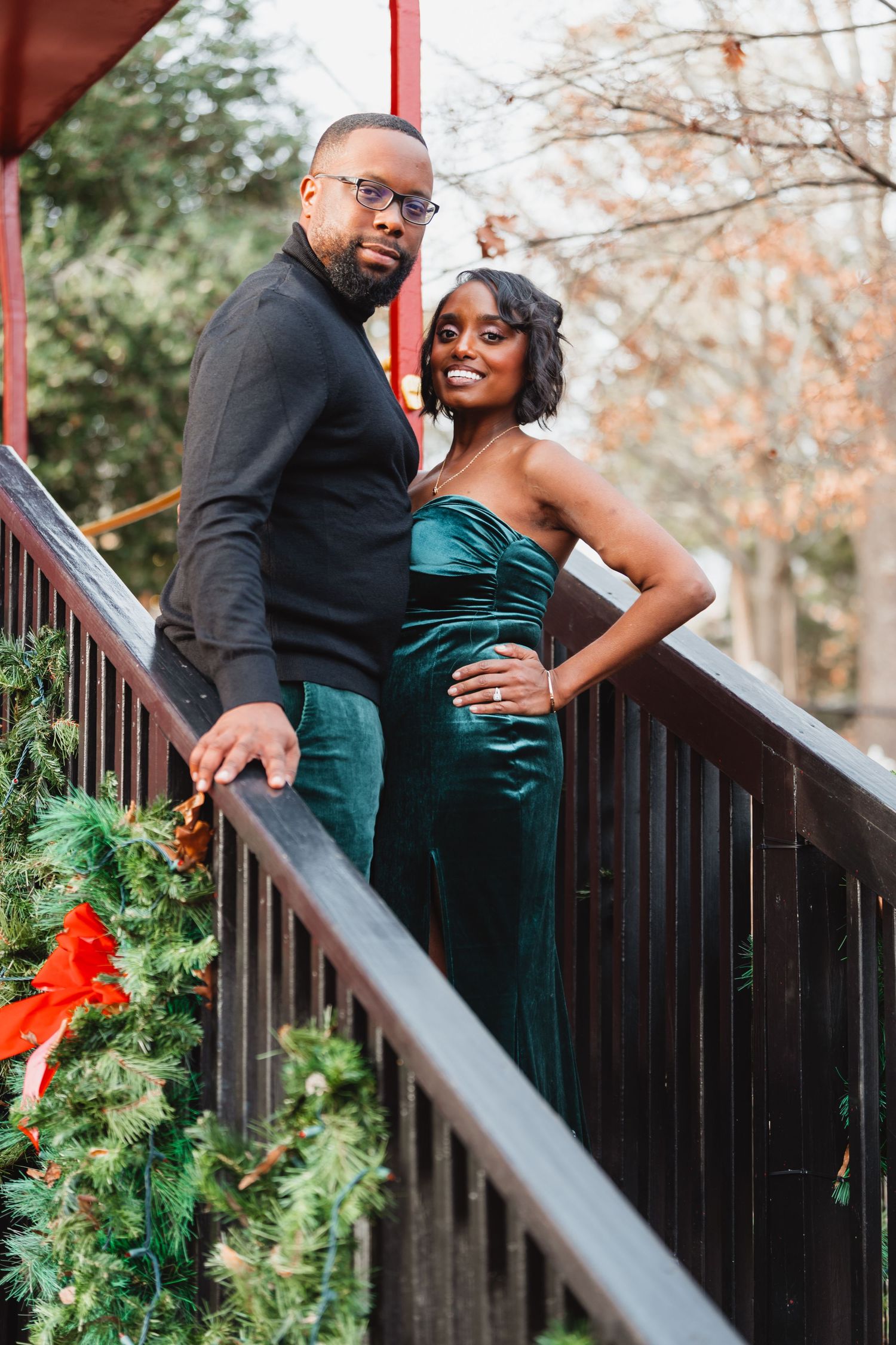 Smiling couple poses on a wooden staircase, the man in black and glasses, the woman in an emerald green dress, near holiday greenery.