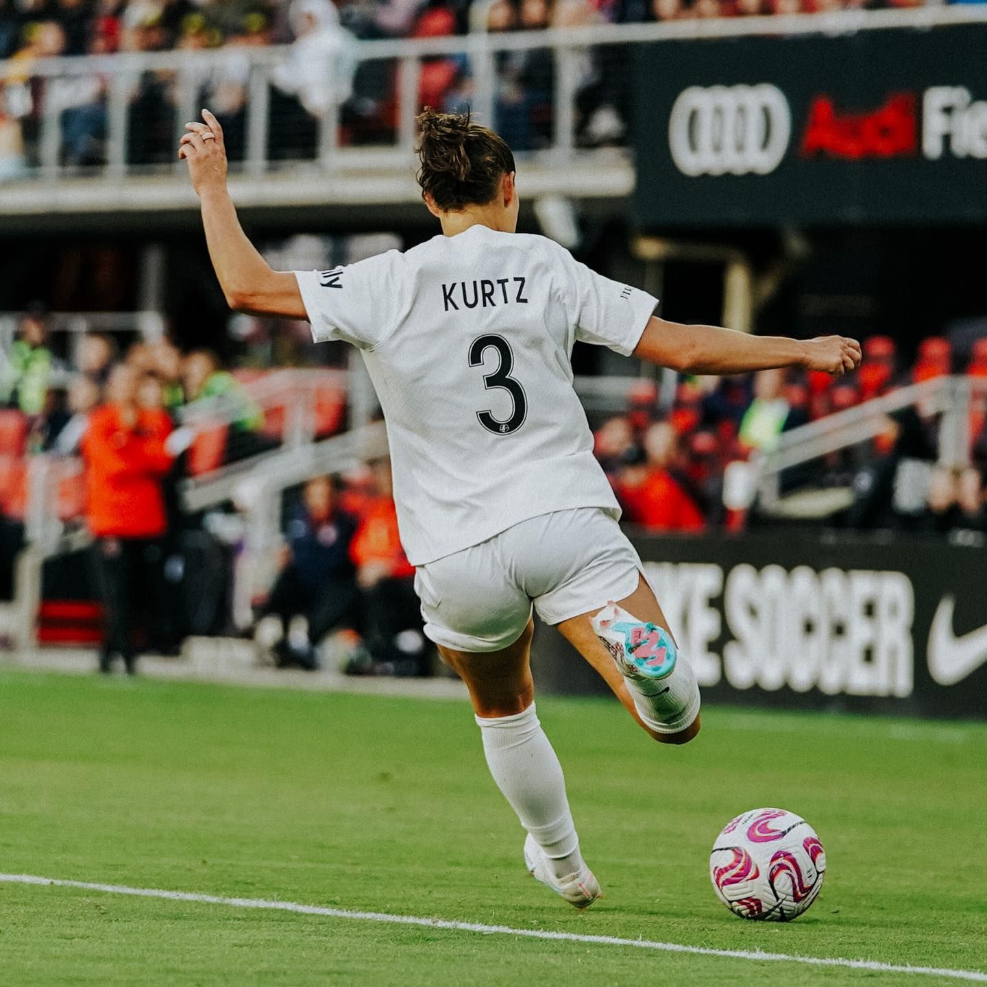 Soccer player number 3 in a white Kurtz jersey strikes the ball on the pitch with fans watching in the stands.