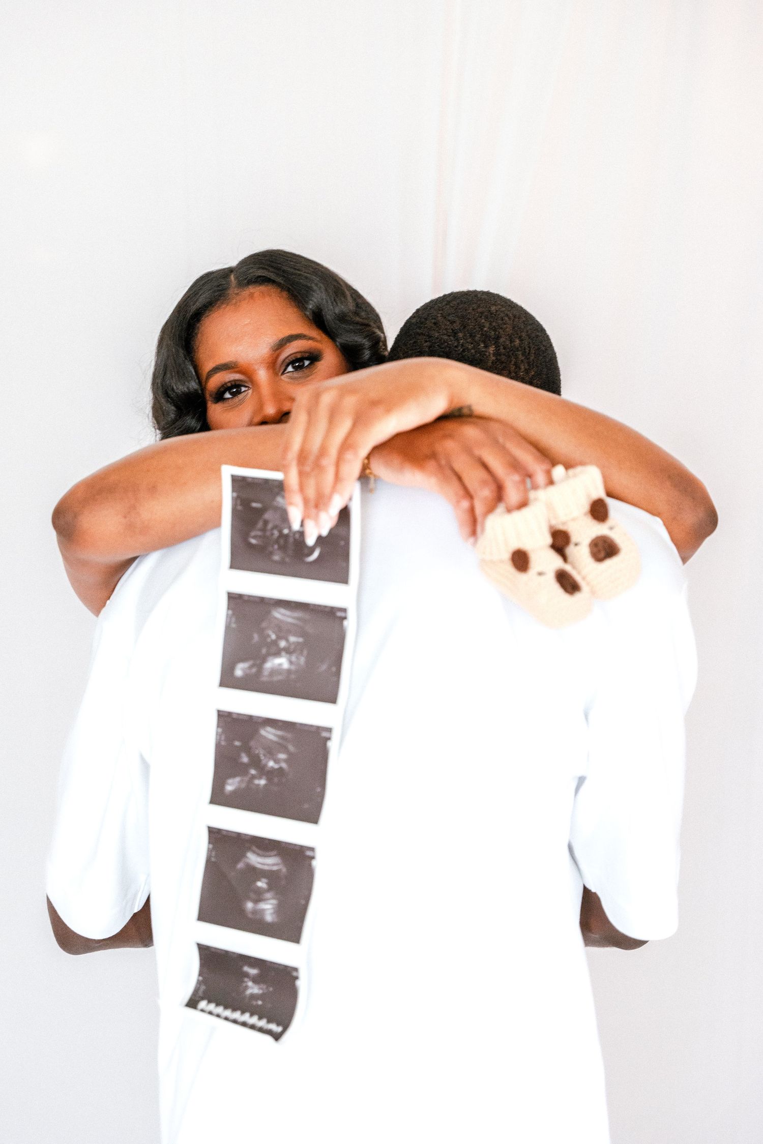 Expectant woman embraces her partner from behind while holding ultrasound photo strips and a small baby shoe in a maternity photo.