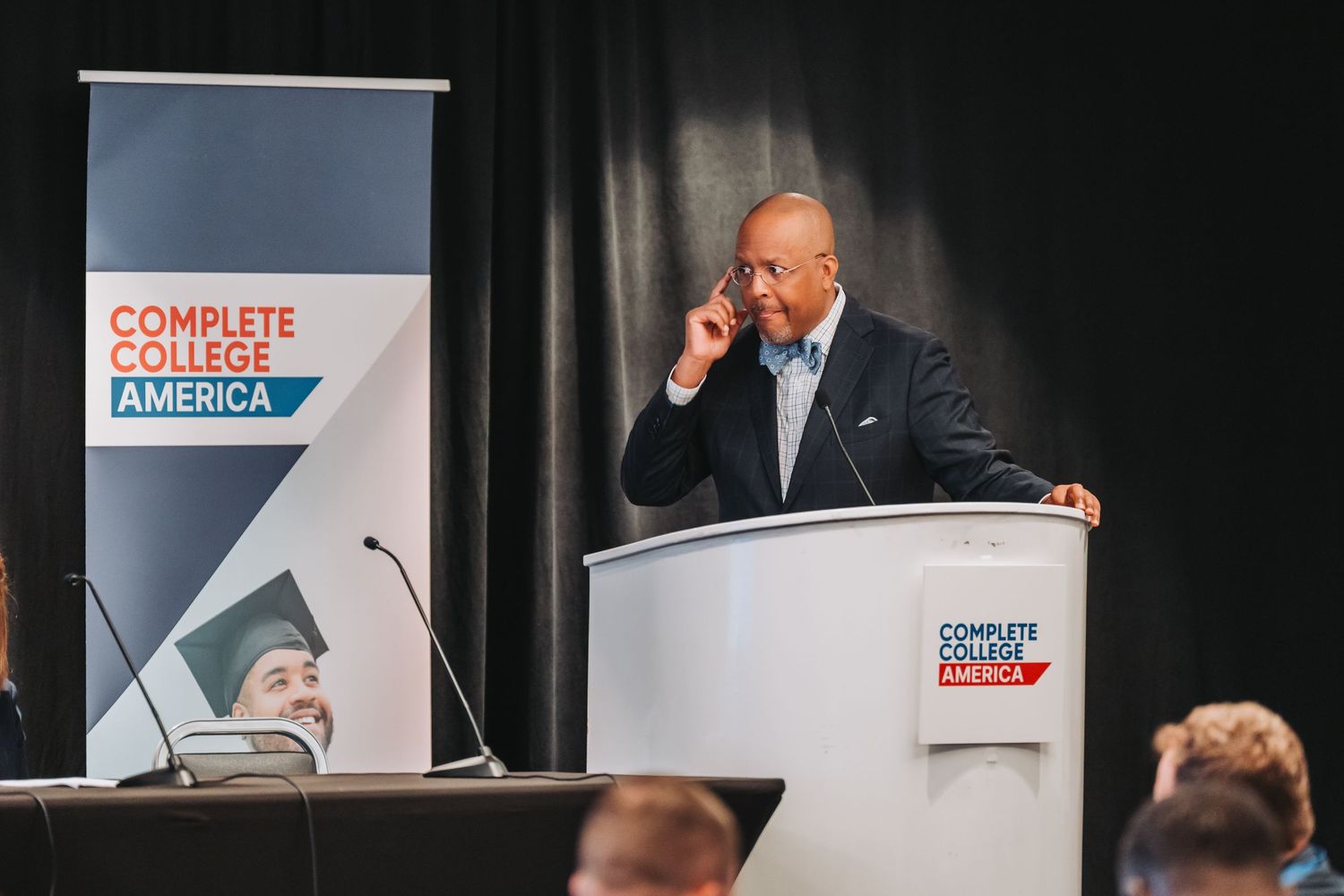 Man in a suit speaks on a phone at a podium during a Complete College America conference, with a banner displayed beside the stage.