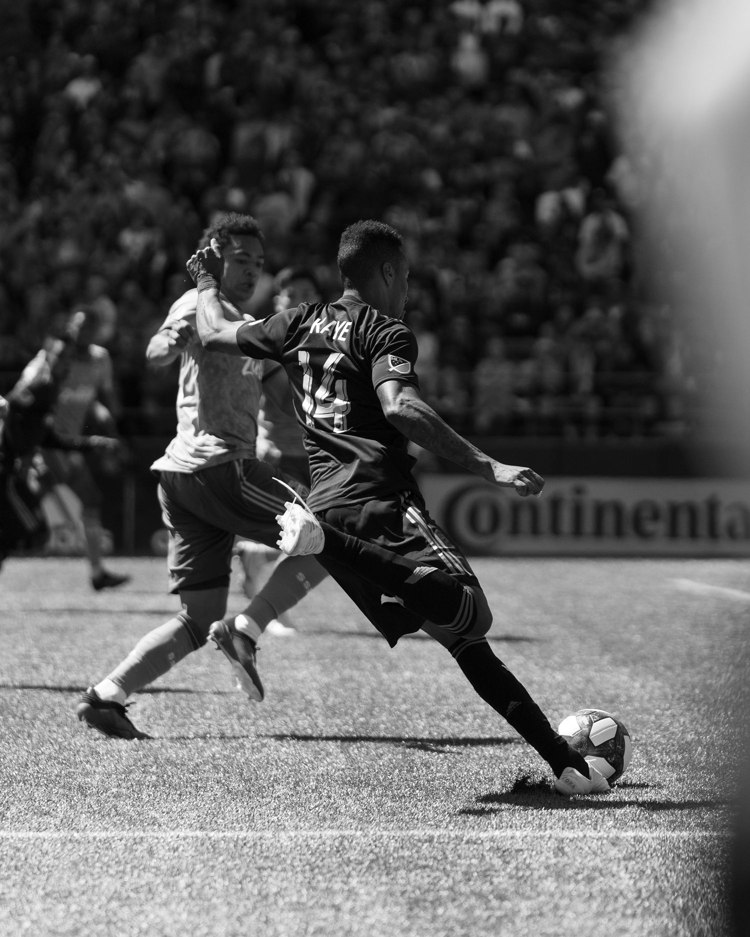 Black and white photo of a soccer player in a dark jersey striking the ball mid-stride while a defender chases during a match.