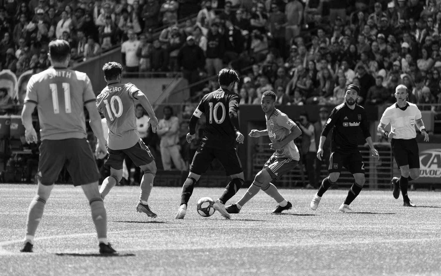 Black and white photo of soccer players in action as a midfielder dribbles the ball past defenders while a referee and crowd look on.