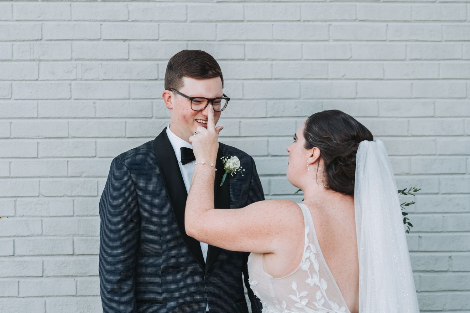 A smiling groom in glasses and a black tuxedo wipes his eye as his bride in a veil touches his face in front of a white brick wall.