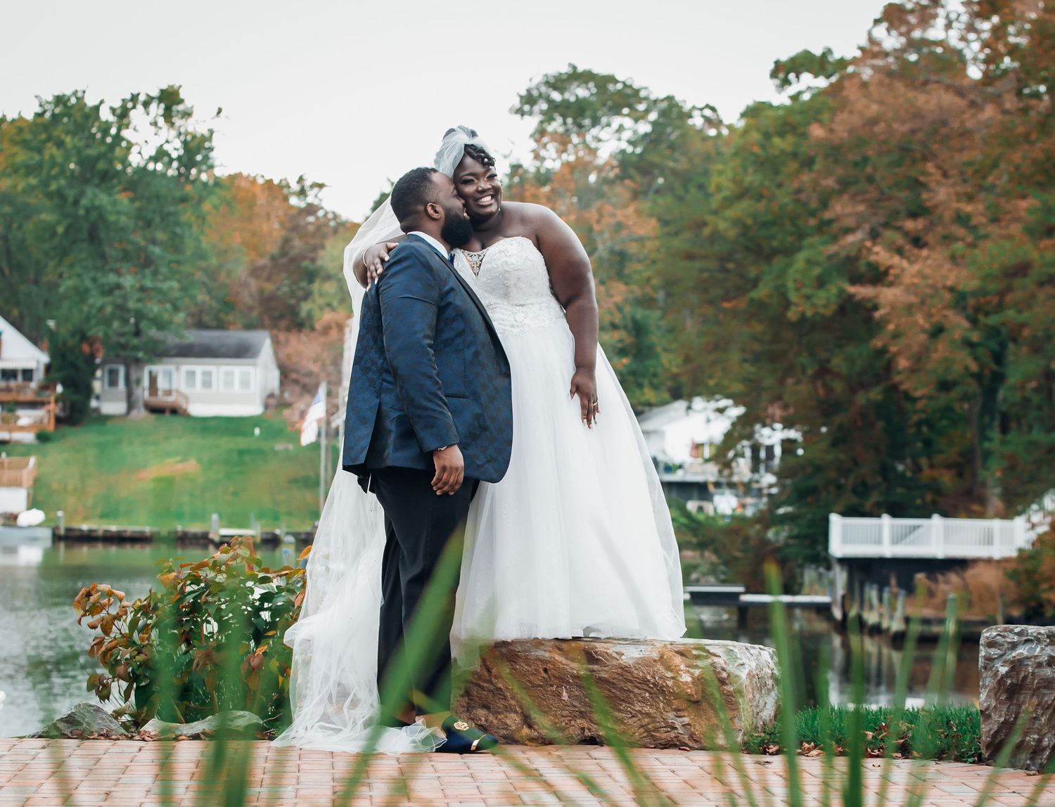 Bride in a white gown and groom in a navy suit share an affectionate moment by a lake framed by colorful autumn trees.