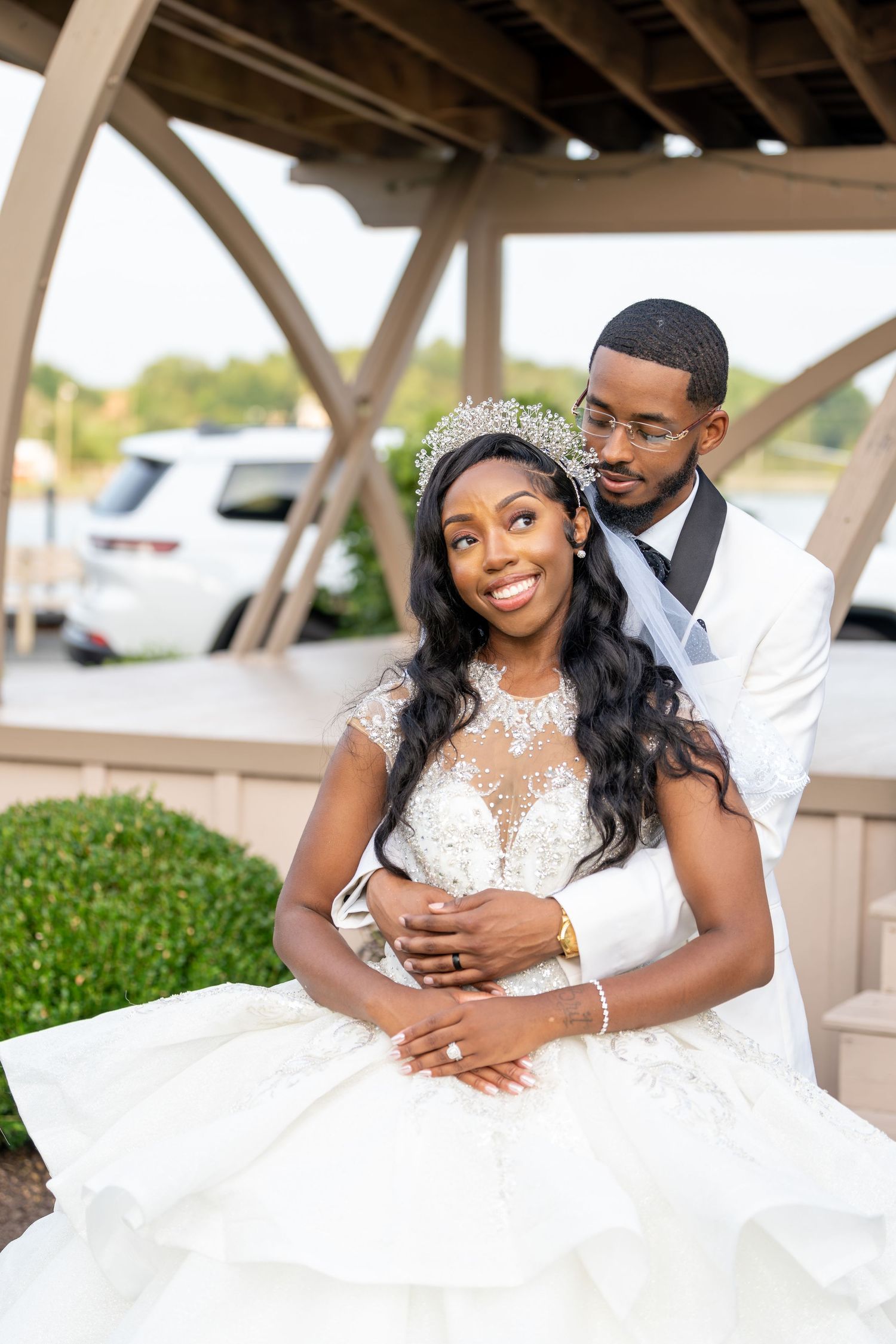 A smiling bride in a sparkling tiara and lace ball gown leans back into her groom in a white jacket beneath a wooden gazebo.