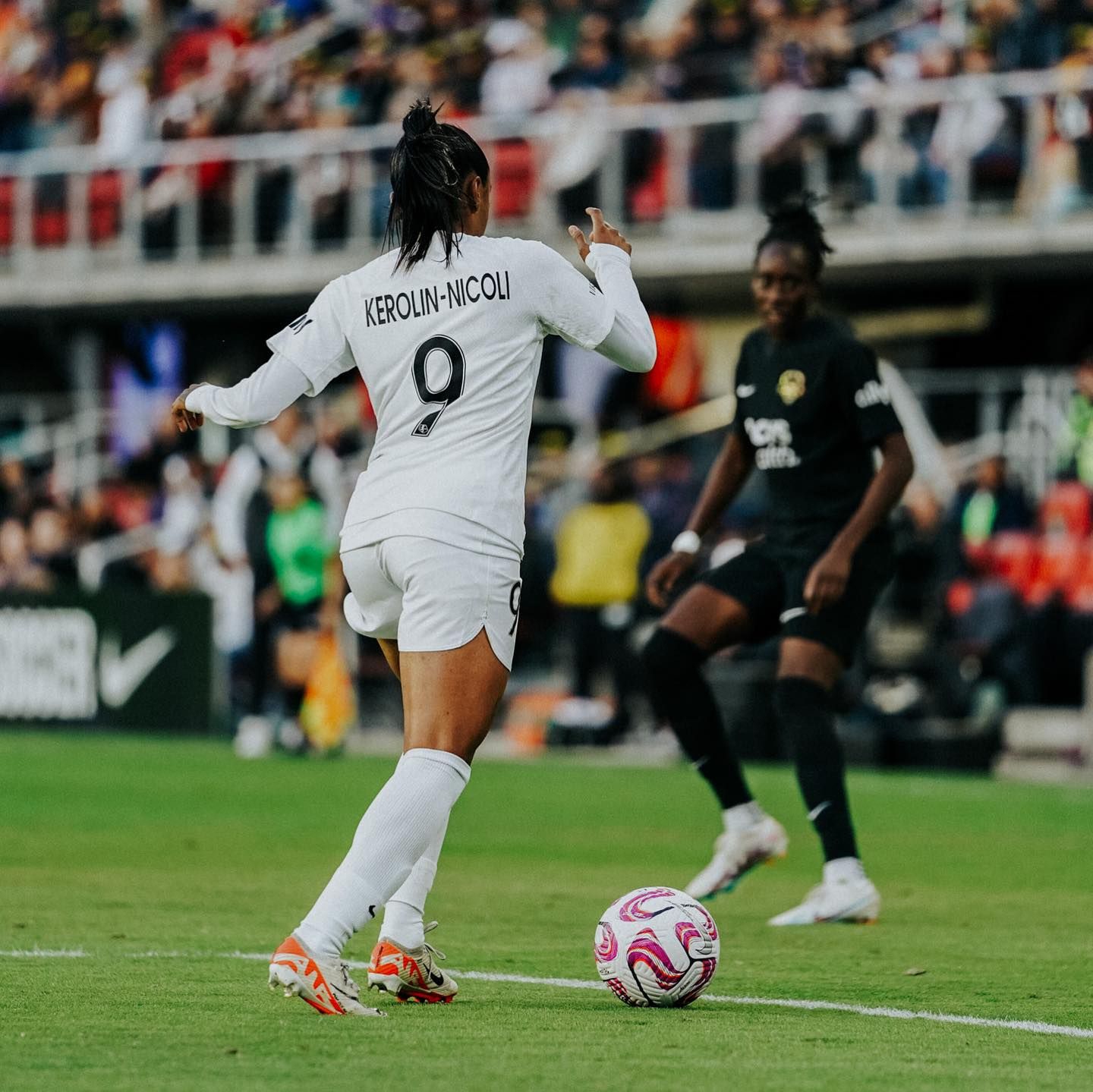 A soccer player in a white number 9 jersey controls the ball on the pitch while an opponent in black looks on.