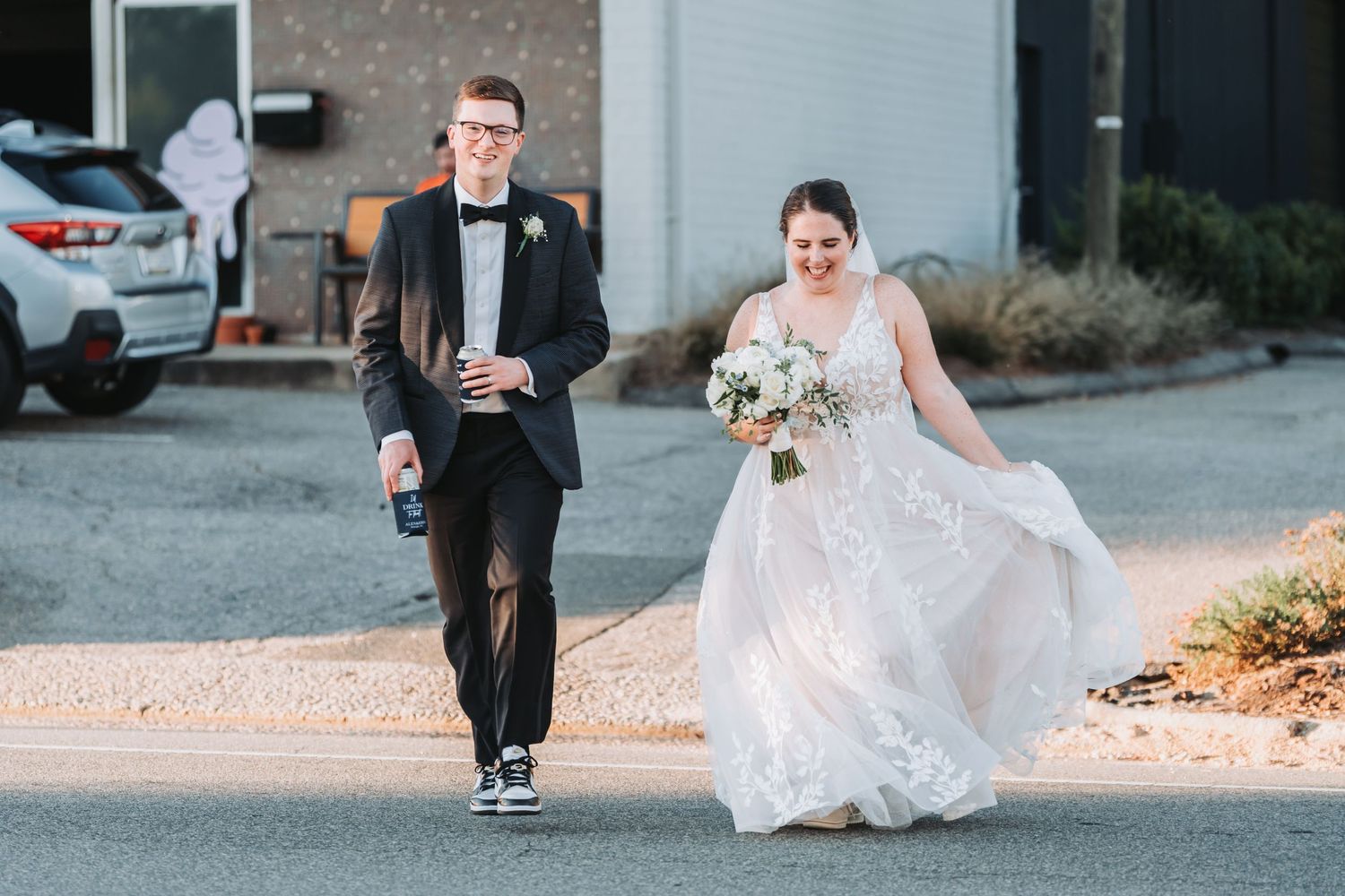 A groom in a black suit and bow tie walks beside his bride, who holds a bouquet and lifts her flowing white gown, down a quiet street.