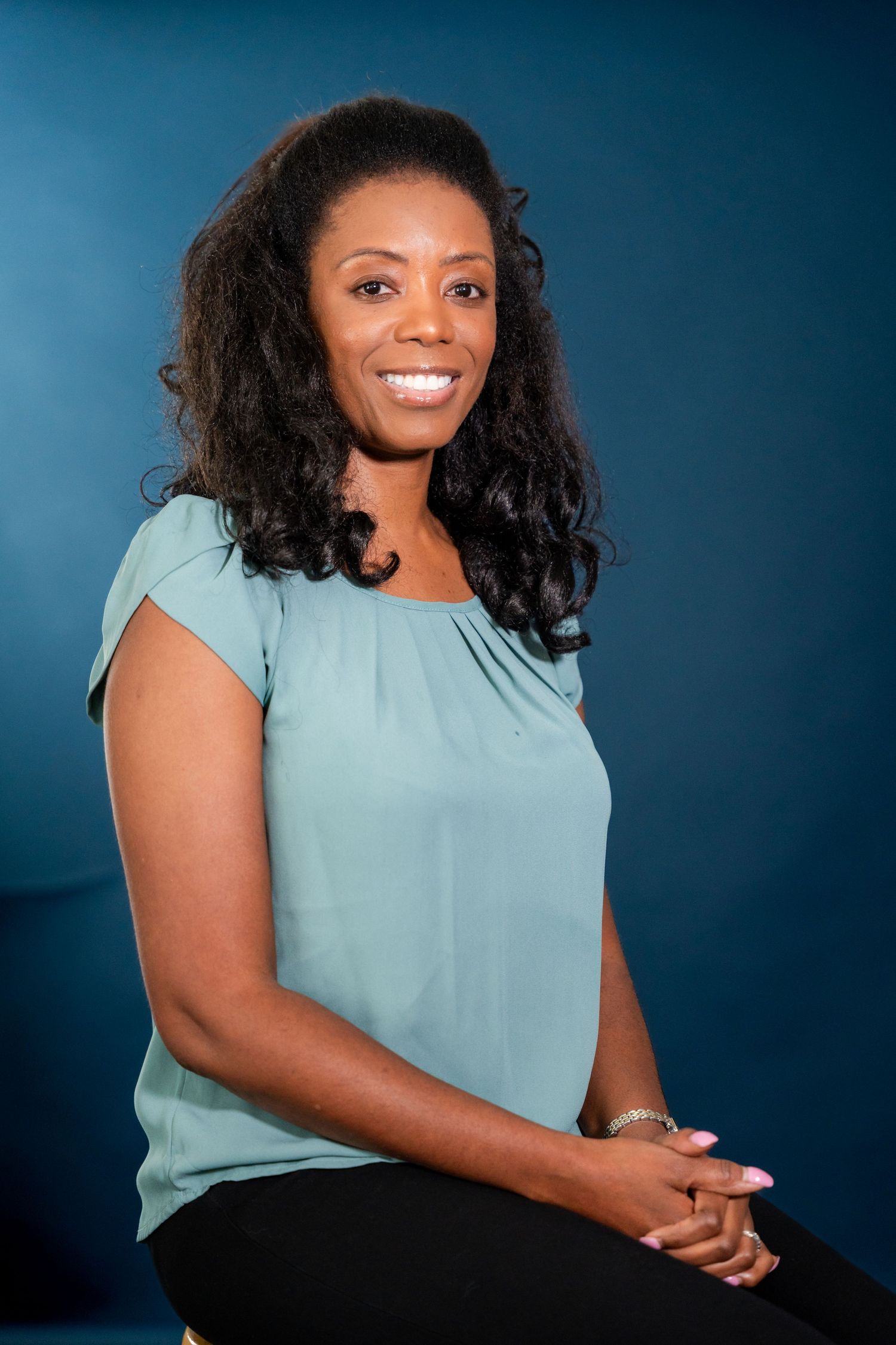 Smiling woman with long curly hair wearing a teal blouse sits against a deep blue studio backdrop for a professional portrait.