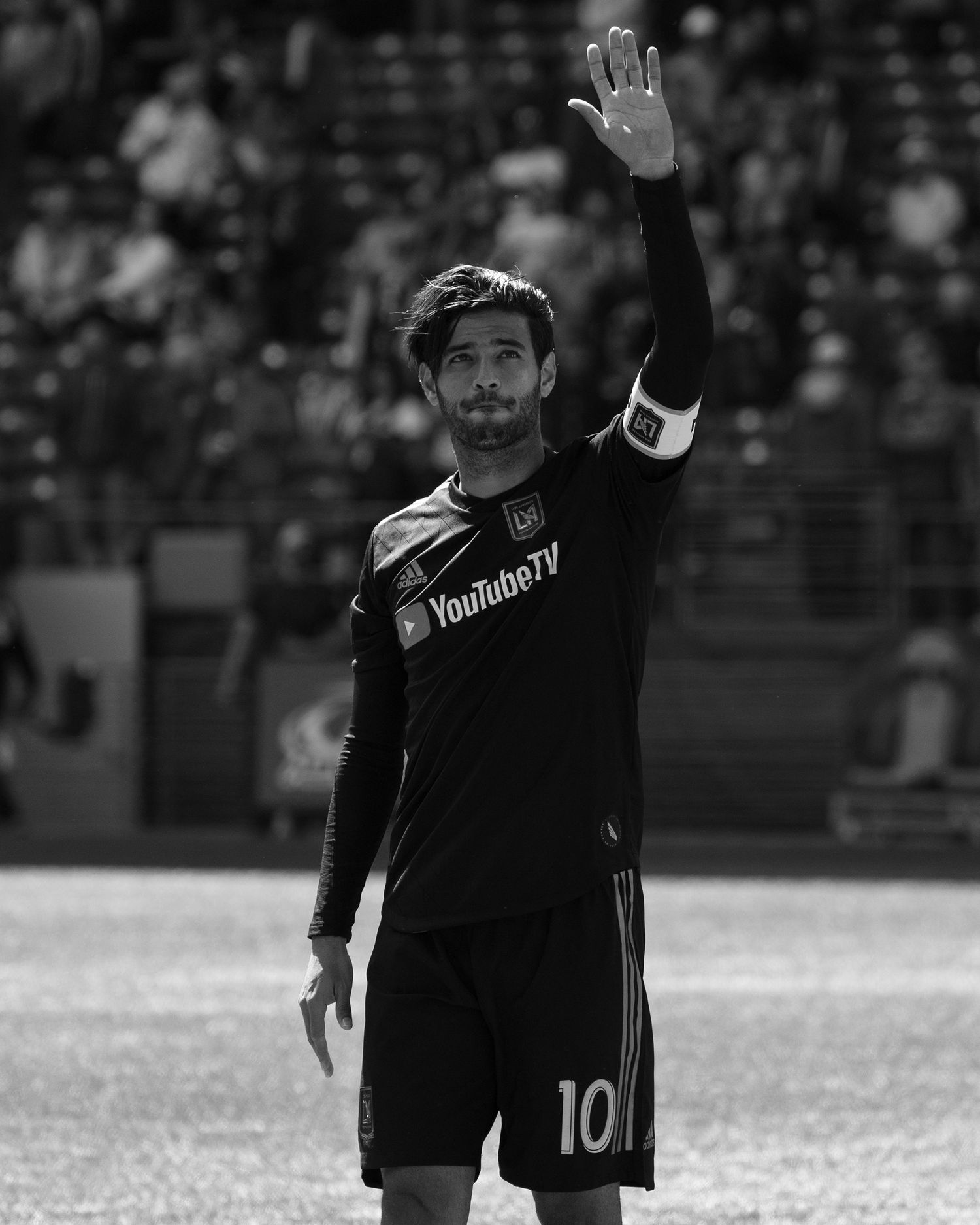 Black and white photo of a soccer player wearing a number 10 jersey and captain's armband raising his right hand on the field.